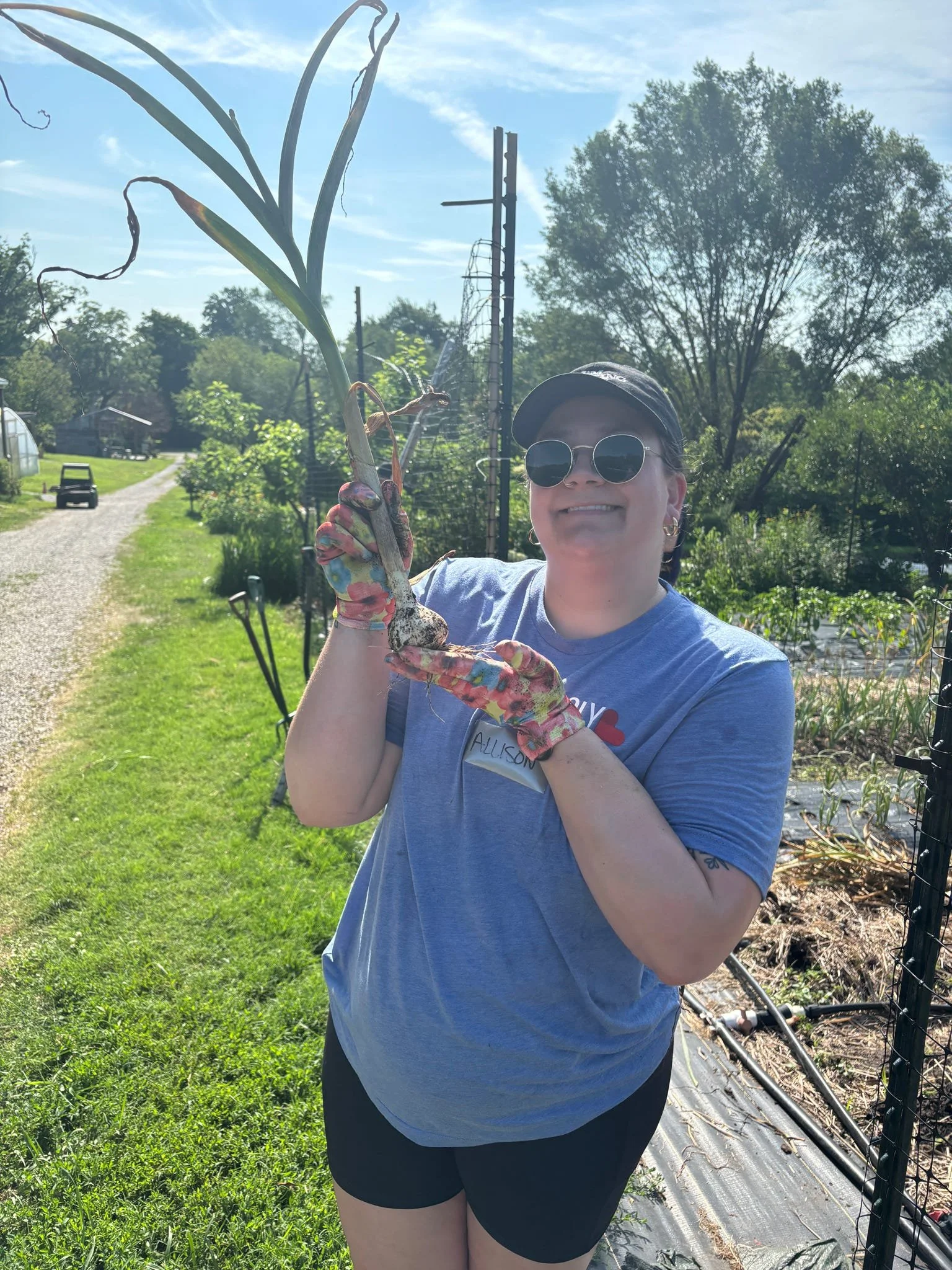 A woman wearing sunglasses, a black cap, a blue t-shirt, and black shorts, smiling and holding a large onion in an outdoor garden or farm.