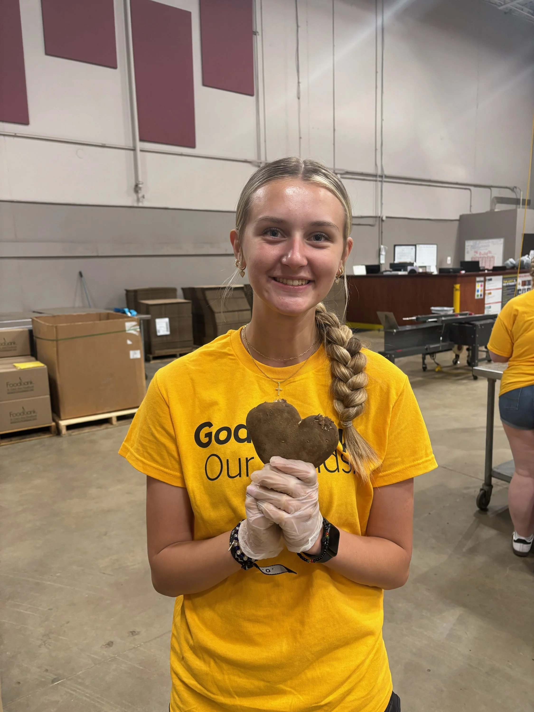 A young woman with blonde hair in a braid, wearing a yellow t-shirt and gloves, holding a large heart-shaped potato and smiling inside a warehouse with boxes and equipment in the background.