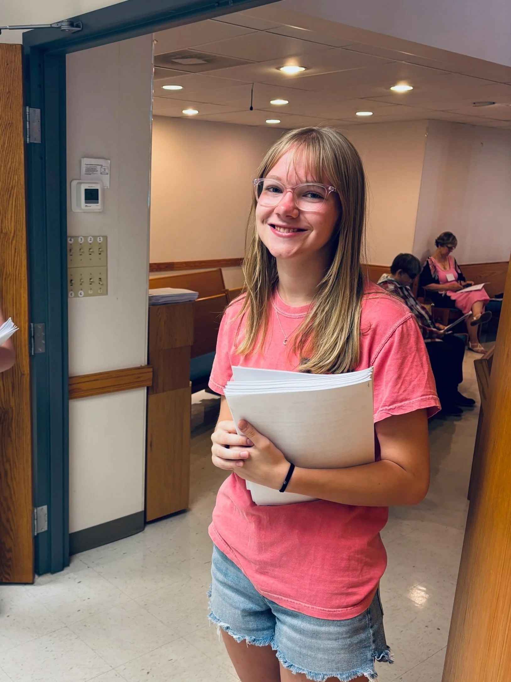 A young woman standing in an indoor setting, smiling, holding a large stack of papers, wearing glasses, a pink T-shirt, and denim shorts.