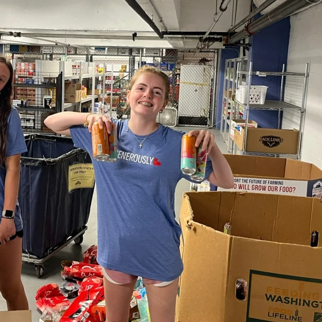A young girl with a big smile, wearing a blue T-shirt and shorts, holding two cans of soda in a warehouse or donation center. Shelves and boxes are visible in the background.