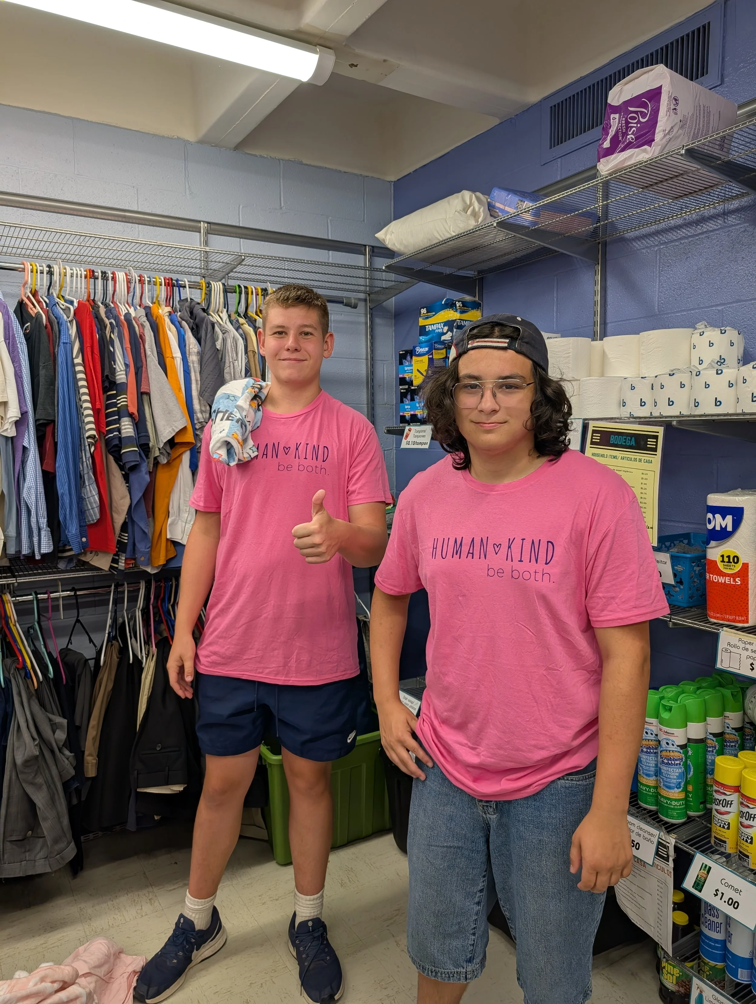 Two teenagers with pink shirts that say 'HUMAN KIND be both.' standing inside a store with clothing and household items. One teenage is giving a thumbs-up, and the other has their hand on their hip.