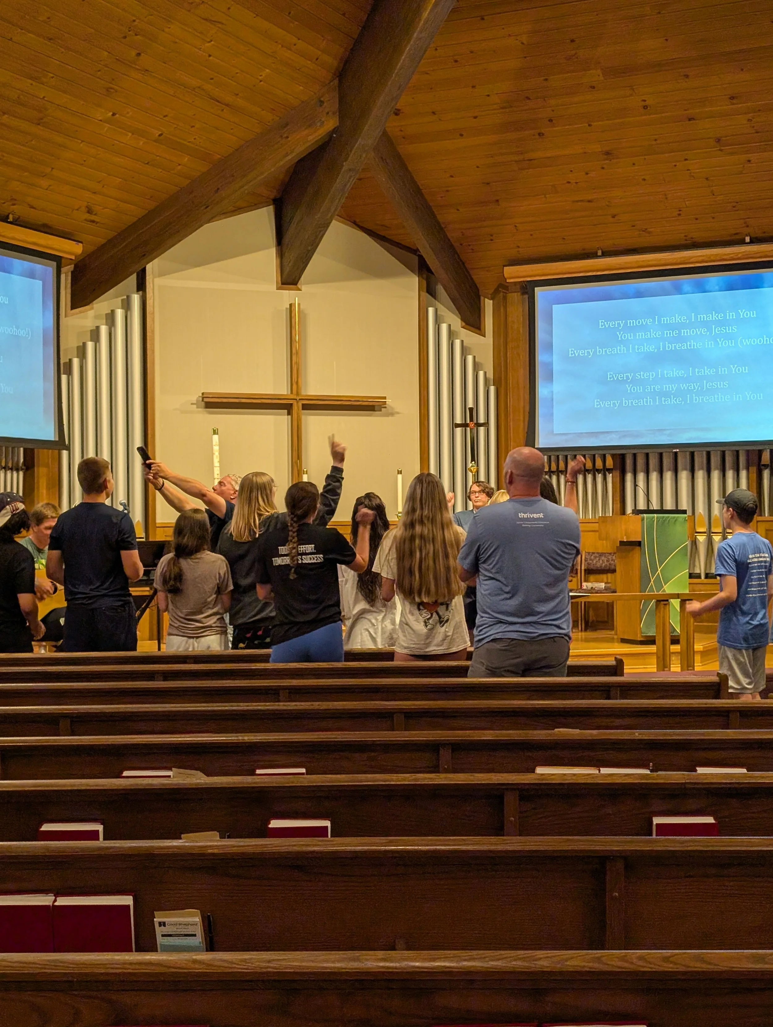 A group of people singing and praising in a church with wooden pews and a large cross at the front. A man is holding a microphone, and there are two large screens displaying lyrics.
