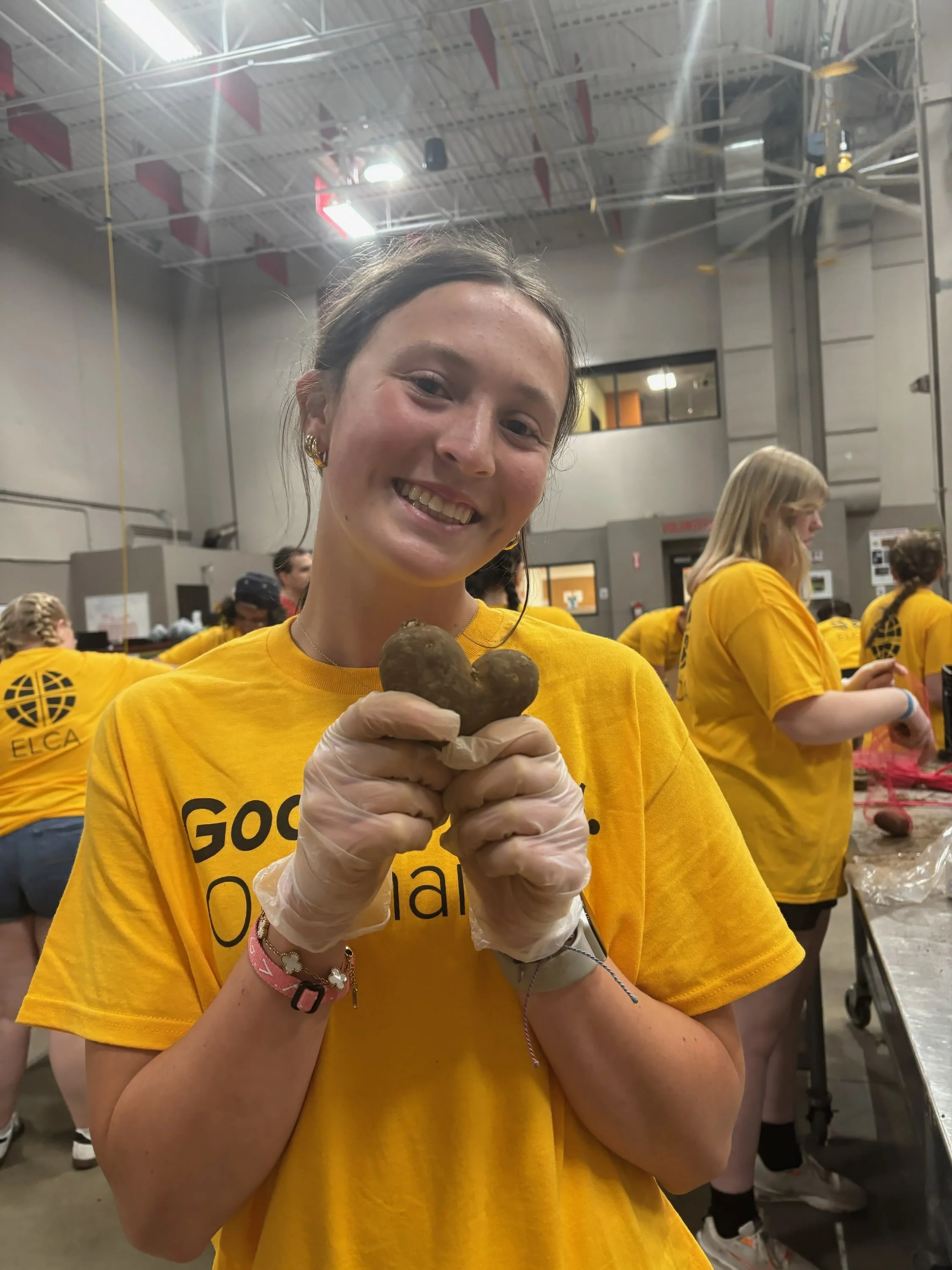 Young woman smiling while holding two small heart-shaped potatoes in a large group activity, all wearing yellow shirts.