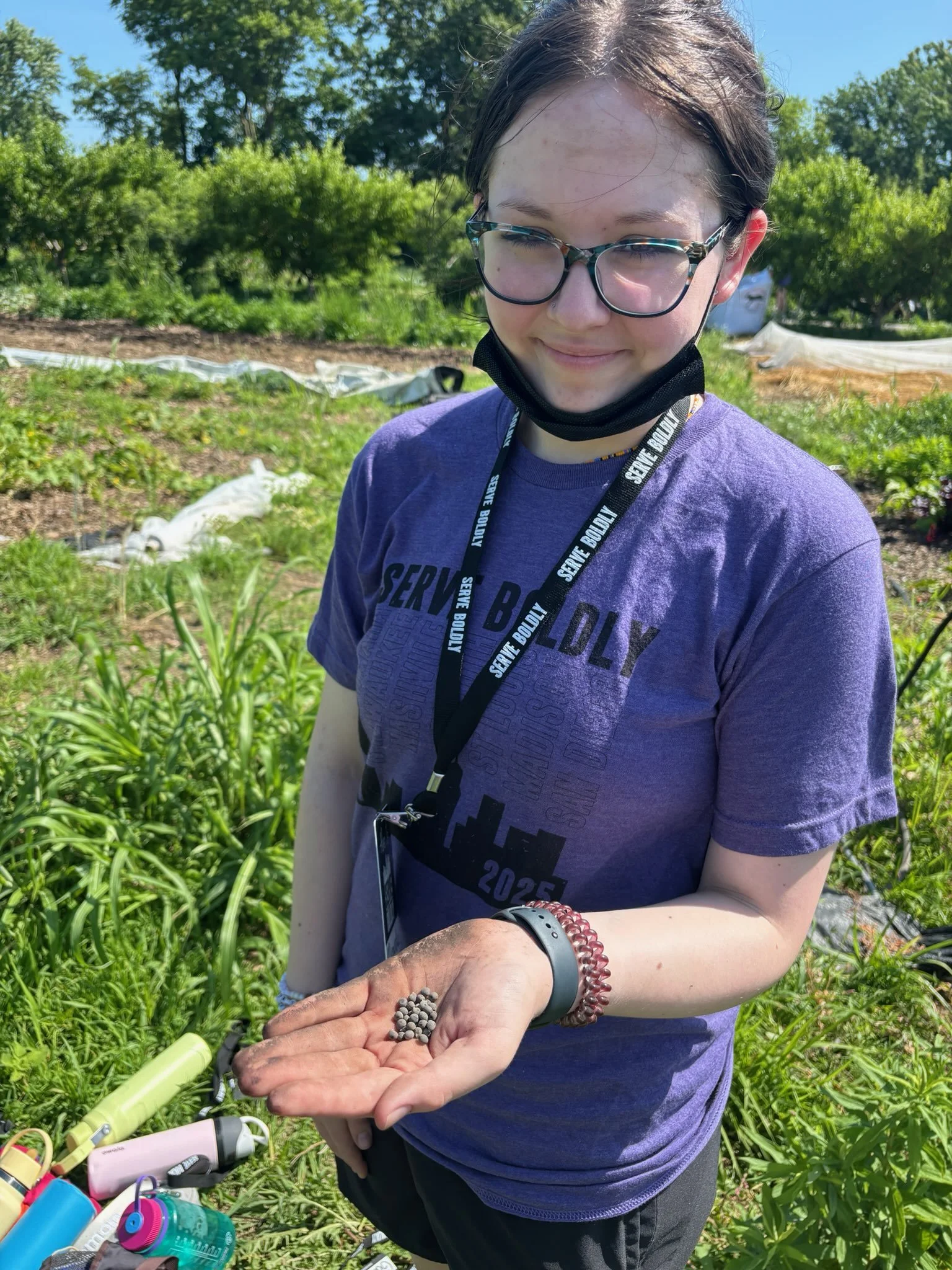 Young girl outdoors in a garden holding a small handful of seeds, wearing glasses, a purple T-shirt, and a black face mask pulled down.