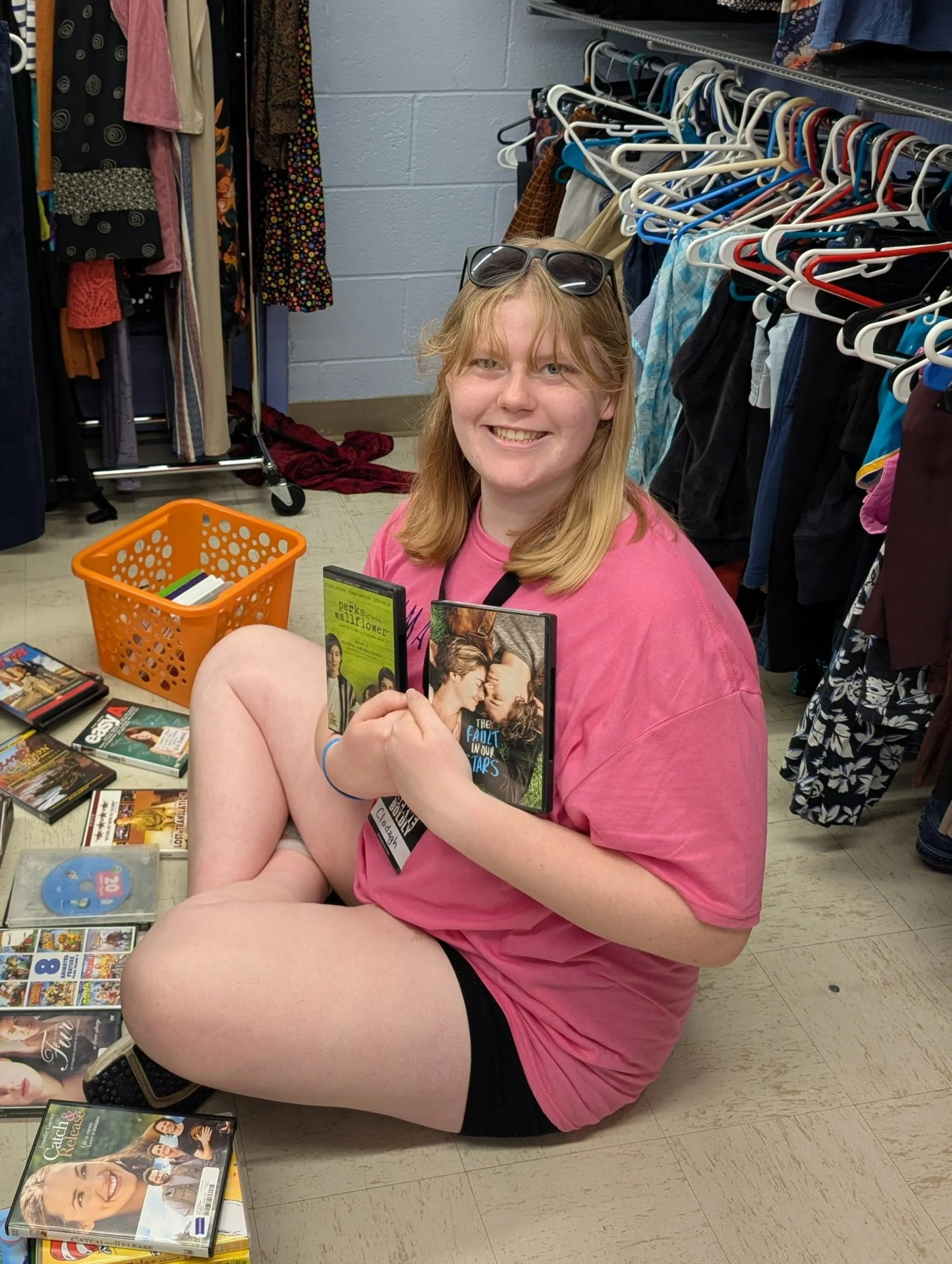 A young woman with blonde hair and sunglasses on her head sitting on the floor at a thrift store. She is wearing a pink T-shirt and black shorts, smiling at the camera while holding two DVDs. There are more DVDs on the floor and clothing racks with various clothes behind her.