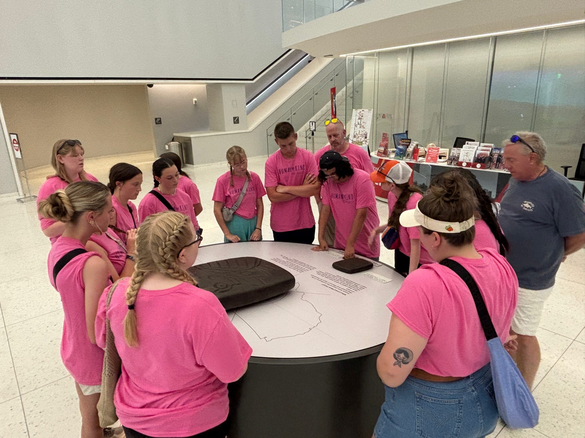 Group of people wearing matching pink shirts gathered around a table with an exhibit, listening to an explanation at a museum.