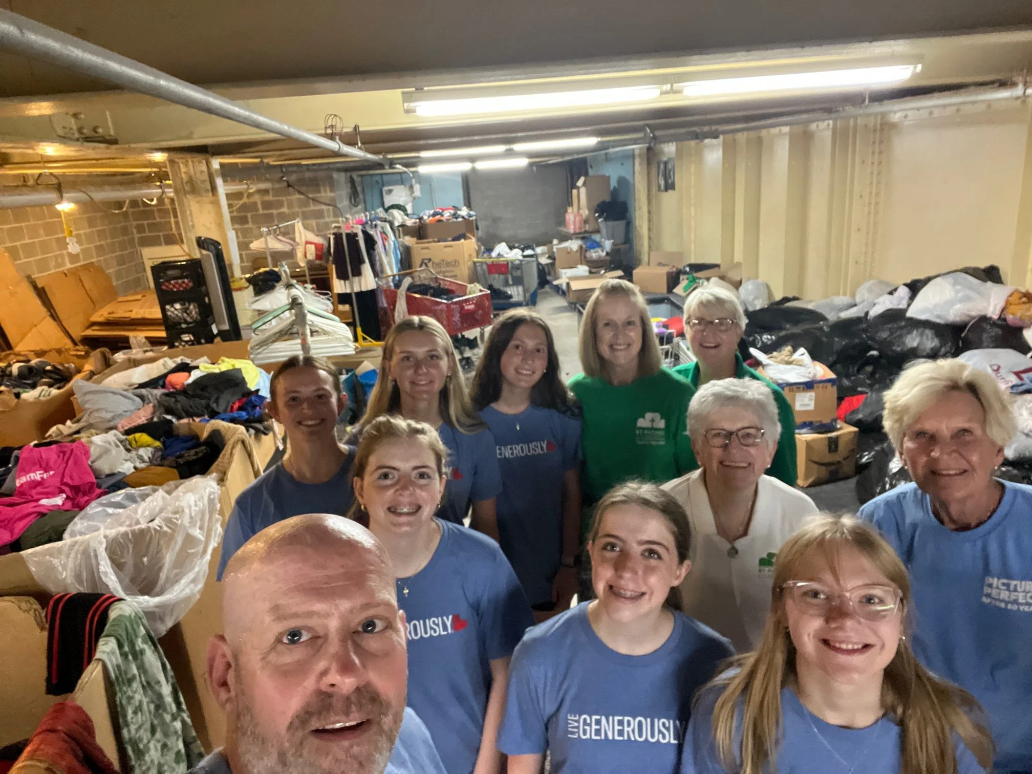 Group of people standing in front of a large pile of clothing and donations in a storage room or warehouse.
