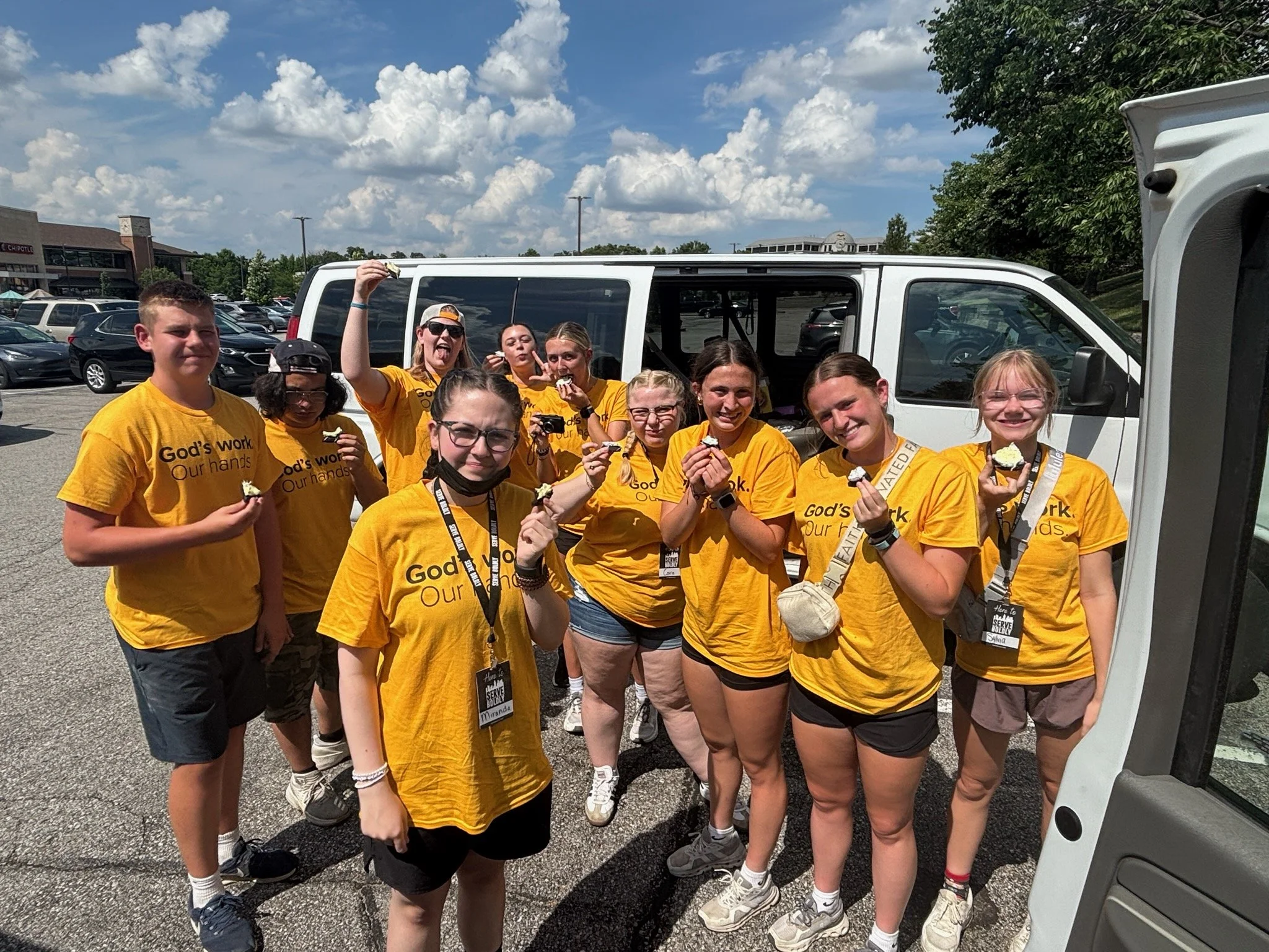 A group of young people in yellow T-shirts standing together outdoors in a parking lot, holding cupcakes and smiling, with a white van in the background.
