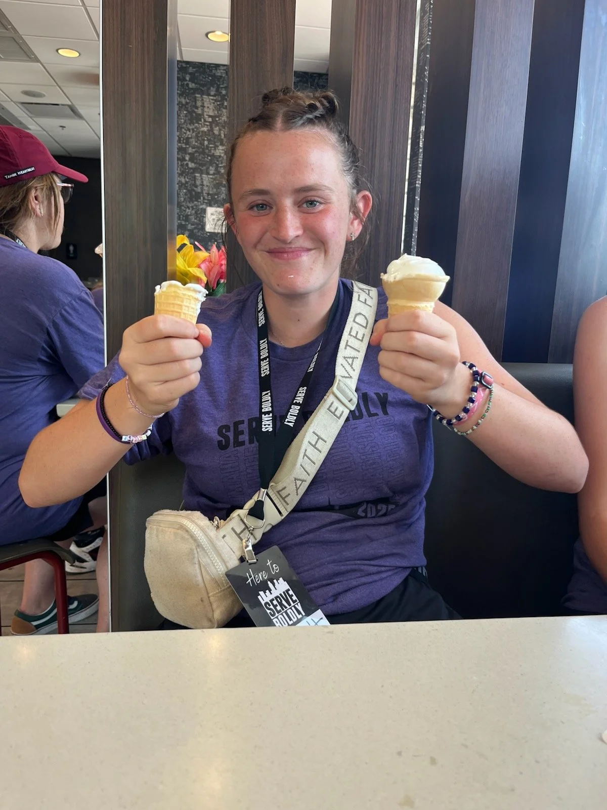A young woman smiling and holding two ice cream cones inside a restaurant or cafe.