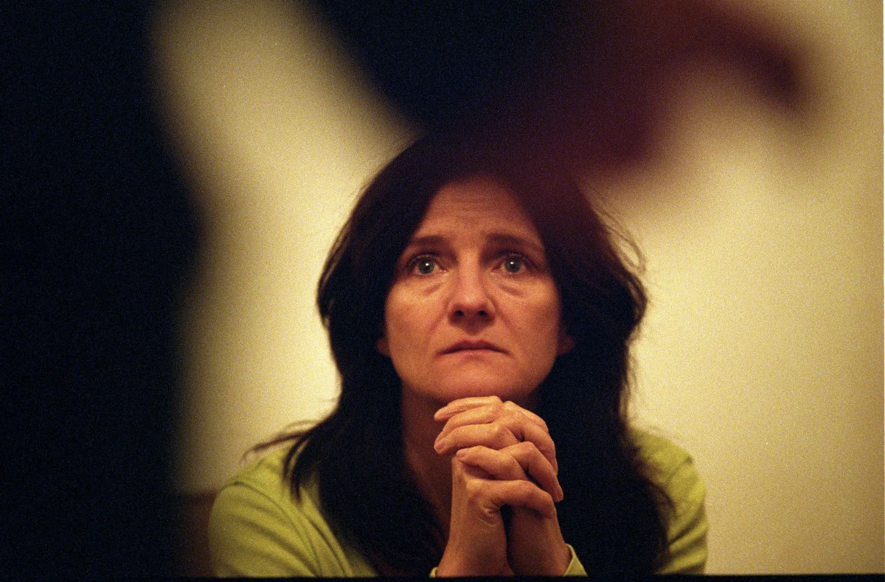 A woman with dark hair and a yellow shirt is sitting with her hands clasped, looking upwards with a serious or contemplative expression.