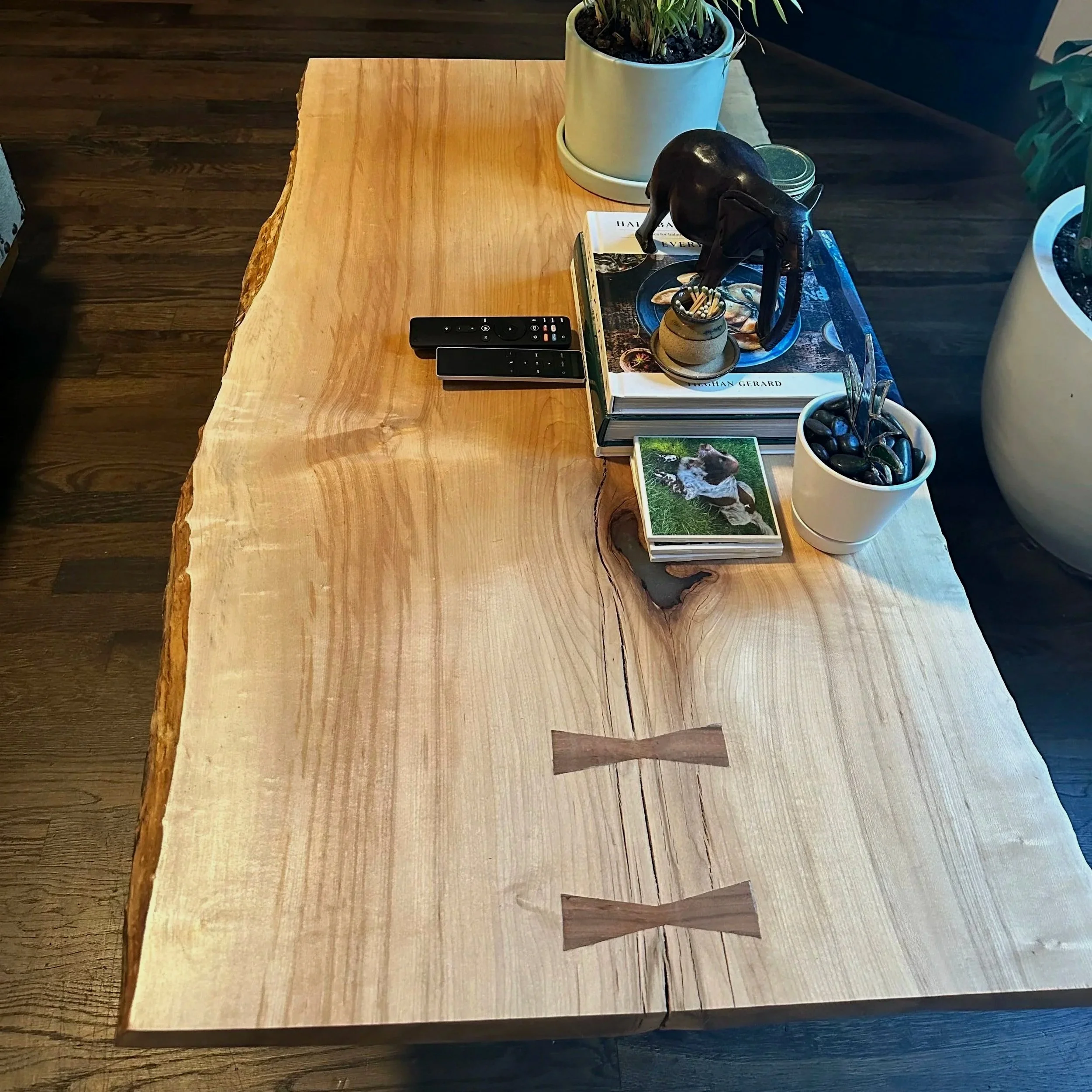 A rustic wooden table with a live edge, decorated with plants, books, remote controls, and decorative items, situated in a room with dark hardwood floors and various potted plants in the background.