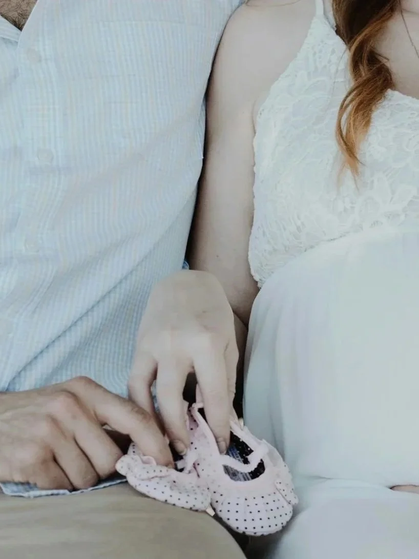 Pregnant white couple sitting together holding baby shoes 
