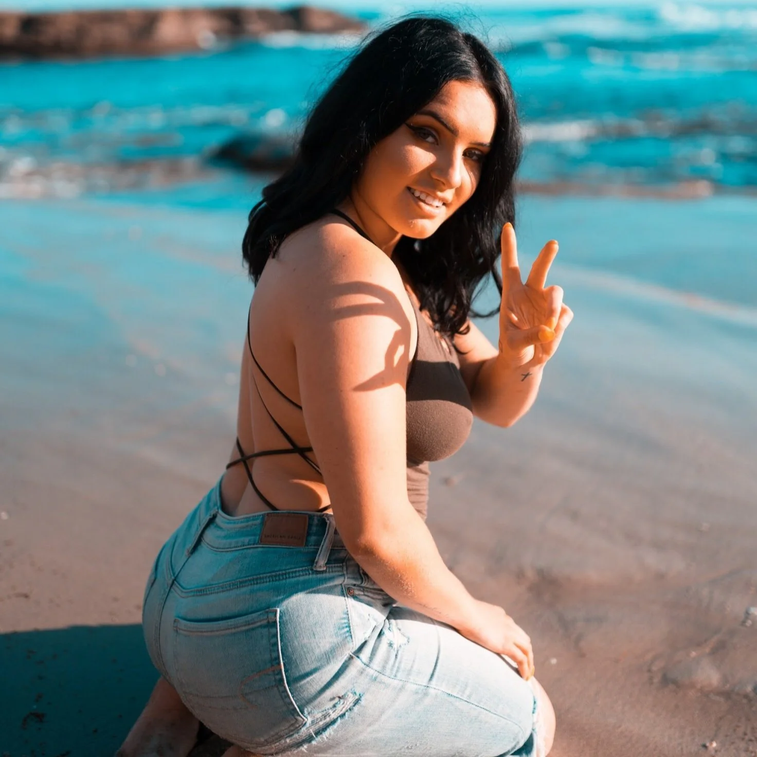 A woman kneeling on a beach, wearing a strappy brown top and jeans, making a peace sign with her hand. The ocean is in the background.