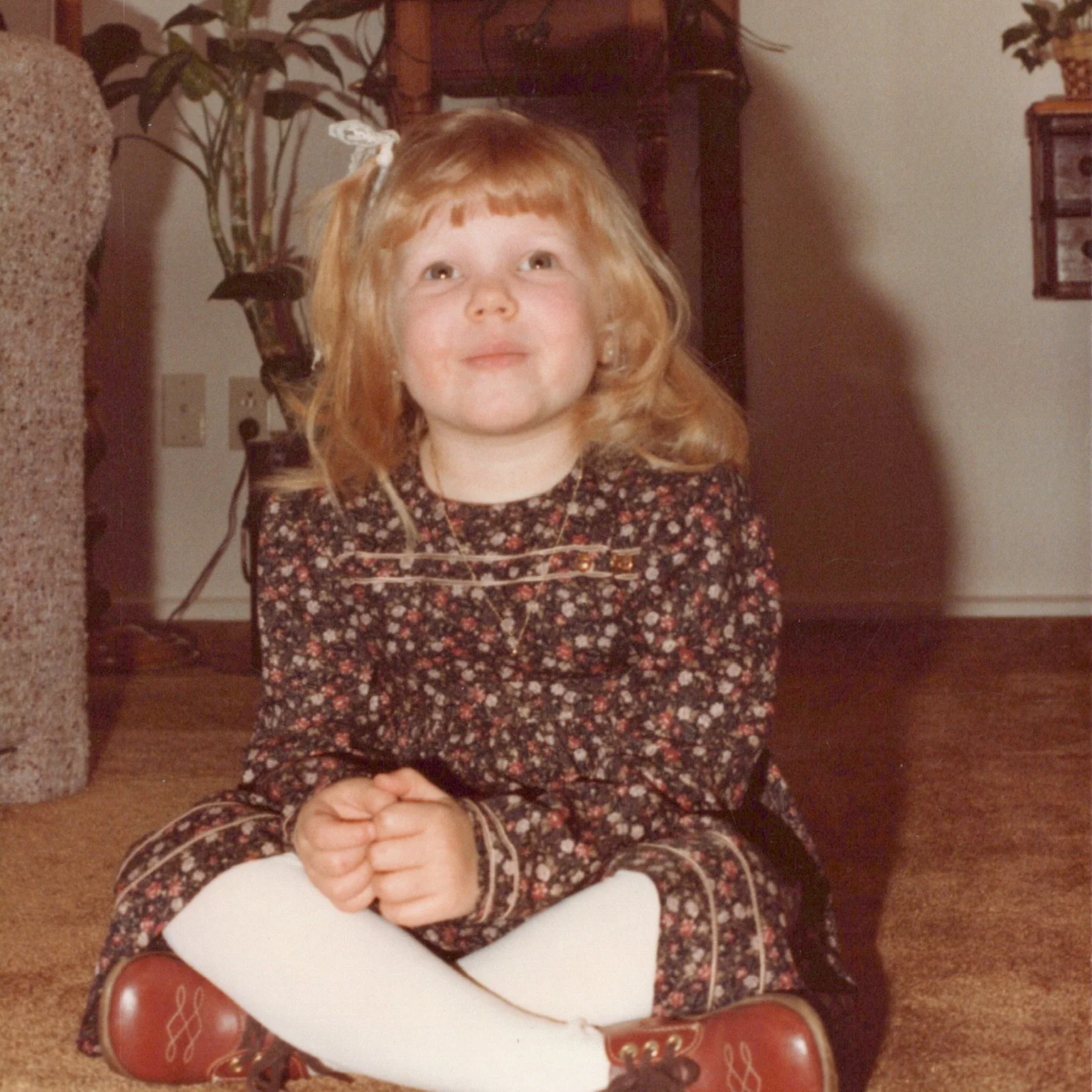 A young girl with red hair sitting on the floor with hands clasped, wearing a floral dress and white tights, inside a home with a plant and furniture in the background.