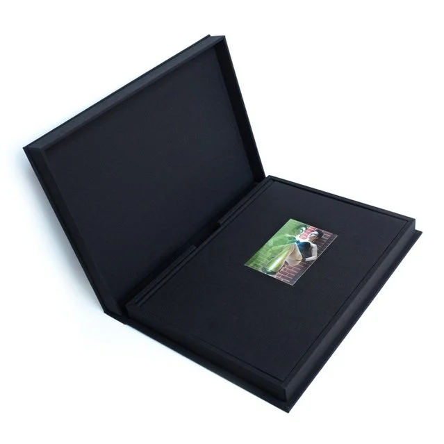 The book clamshell is sitting open. It is wrapped in black fabric. Inside it is a book in black with a small photo in the top center. It shows a woman with black hair wearing black lingerie and heels looking at her reflection. Her leg is held up.