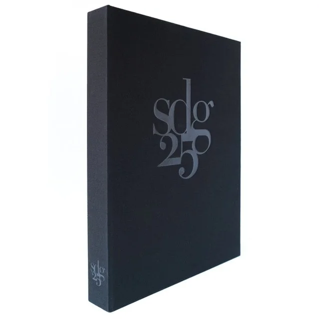 This book clamshell is covered in black fabric. The front has a debased logo with the letters sdg and the numbers 25 interlinked. The logo is also on the spine at the bottom.