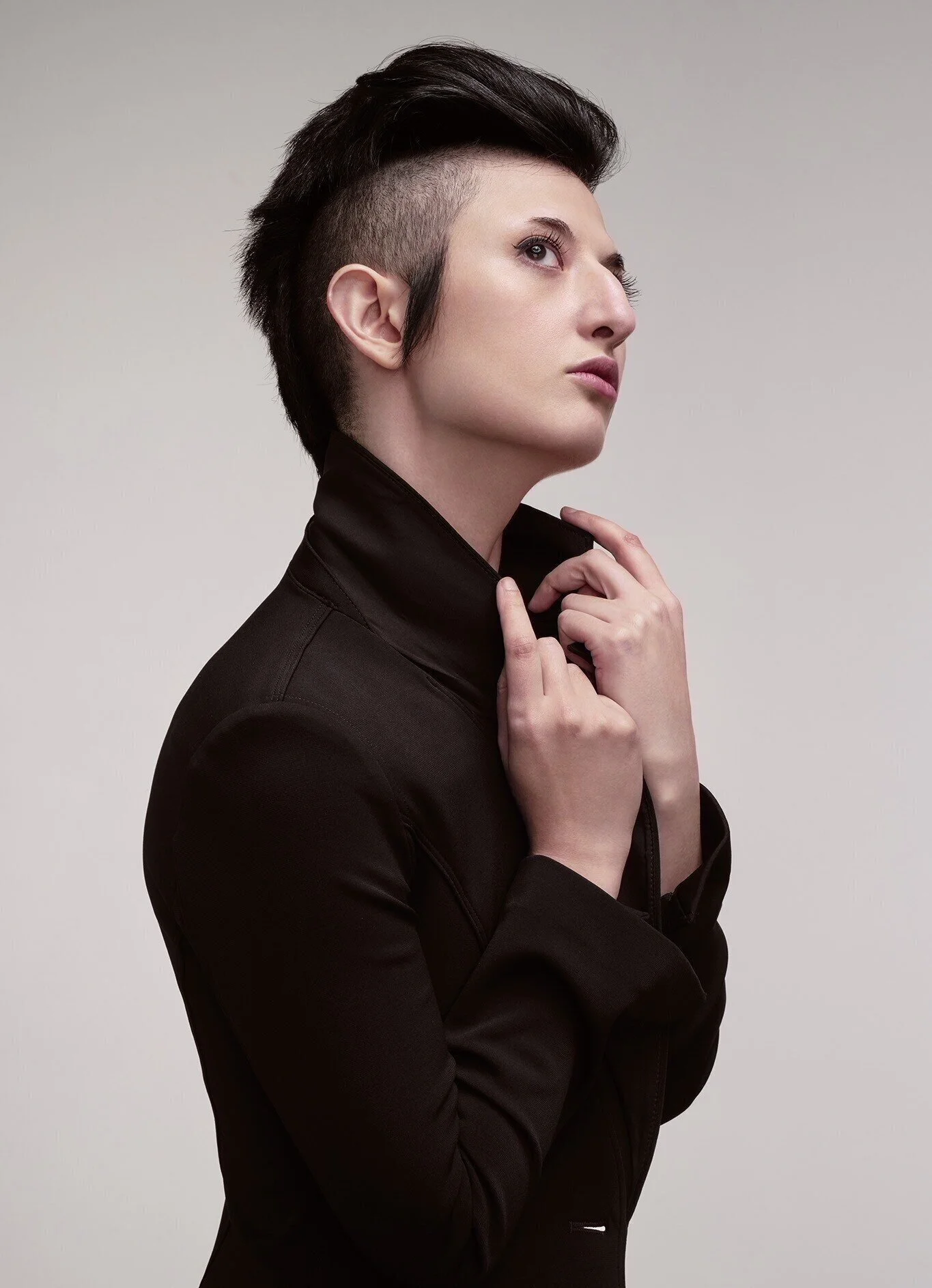 A photo of a woman with a black mohawk facing the right. She is wearing a black blazer and her hands are holding her blazer collar.