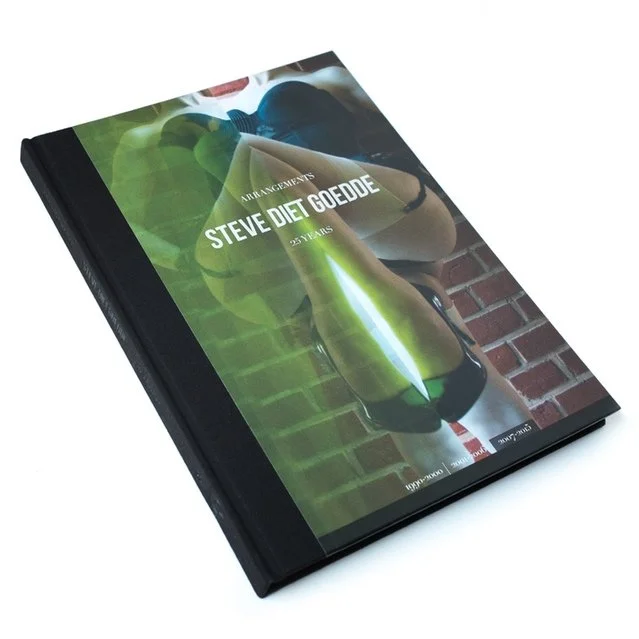This book cover reads "Arrangements" below that "Steve Diet Goedde" below that "25 years" On the very bottom is highlights the dates 2007-2015. The cover art is a photo of a woman from the neck down in black lingerie & black patent leather heels.