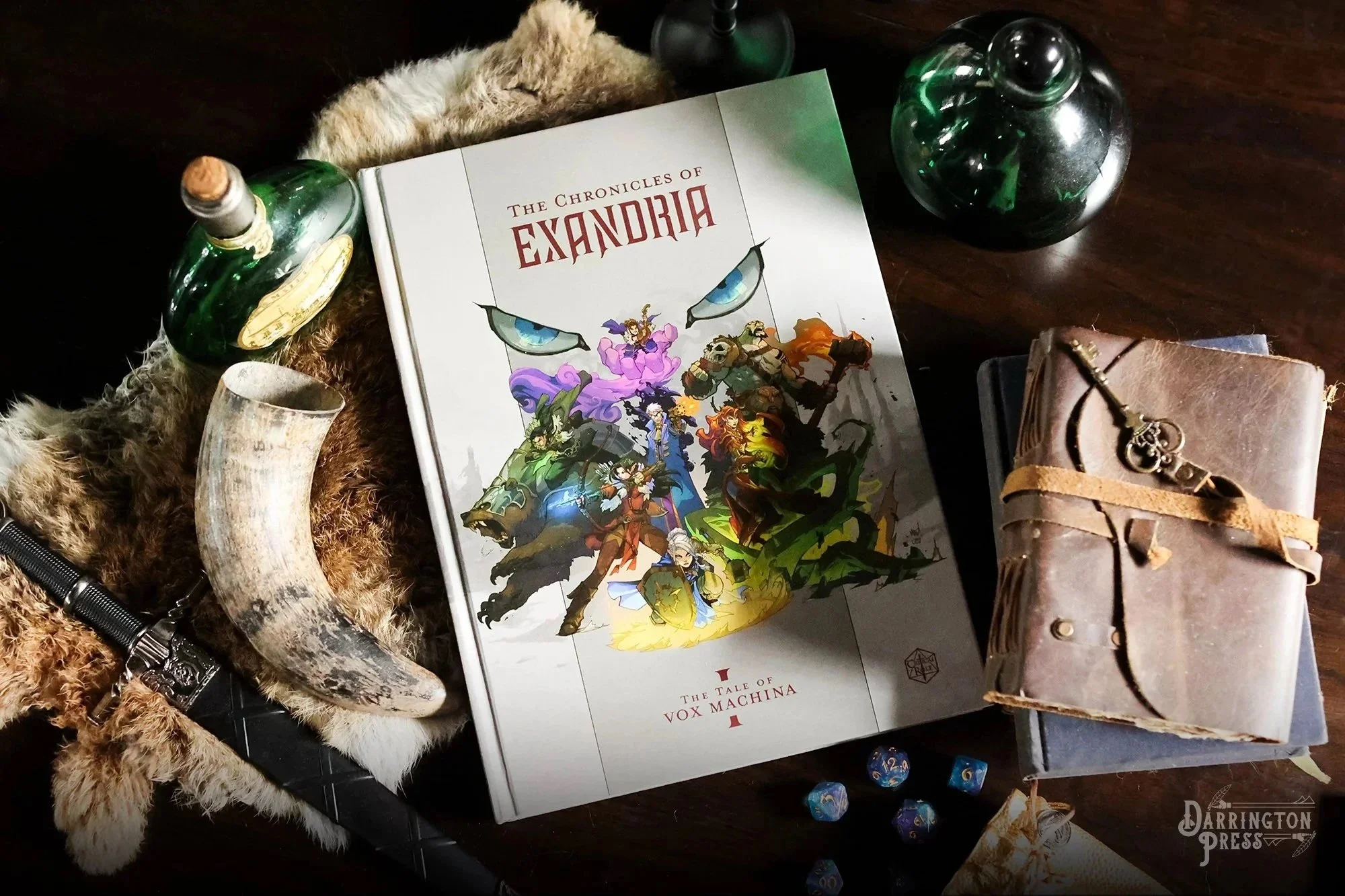 Standard edition of Critical Role: The Chronicles of Exandria Vol. I: The Tale of Vox Machina, showing a fantasy adventuring party (Vox Machina) beneath enormous blue eyes, displayed with dice, potion bottles, a horn, and a leather journal.
