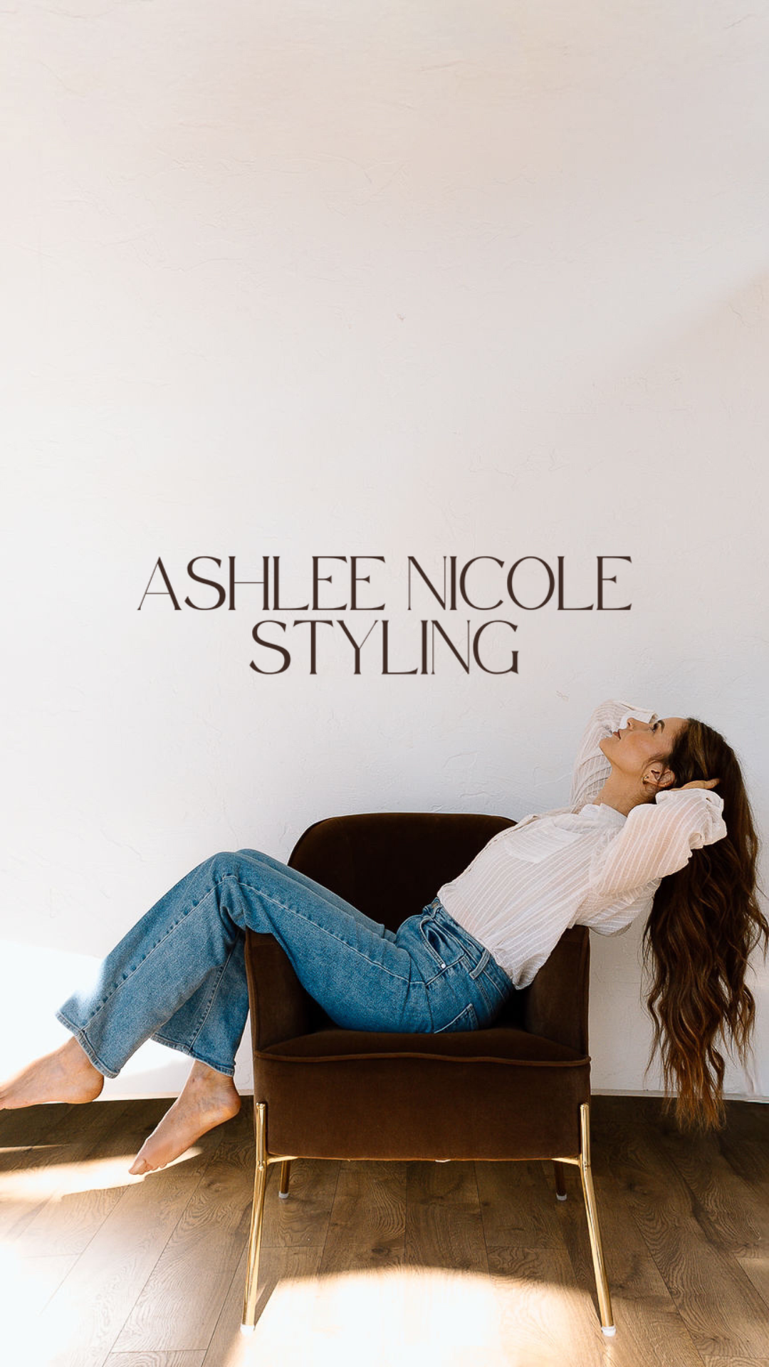 A woman with long brown hair is reclining on a dark brown modern chair, leaning back with her hands behind her head, wearing a white blouse and blue jeans, against a plain light-colored wall.