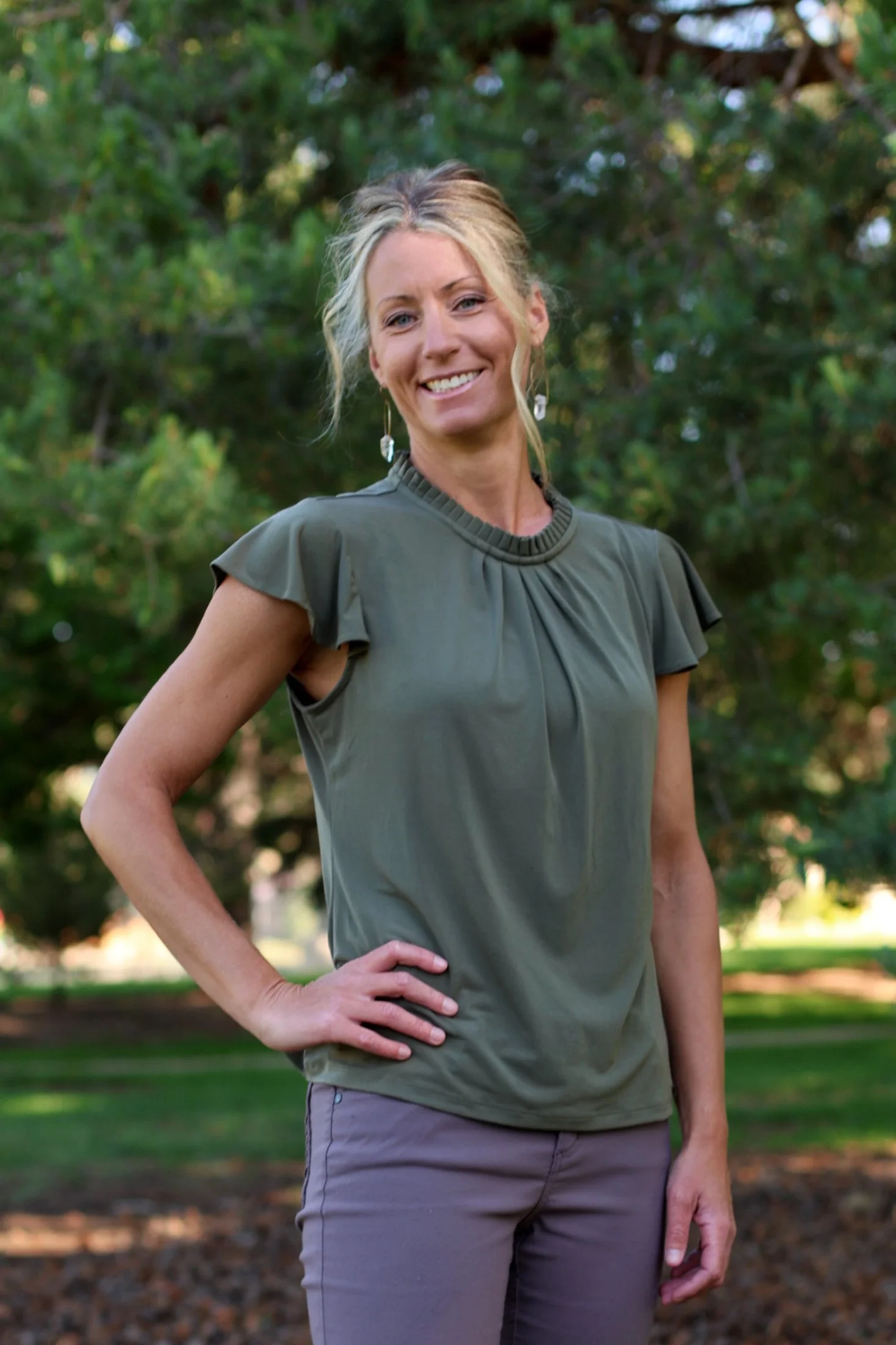 A woman with blond hair and blue eyes smiling outdoors near green pine trees, wearing a cream-colored long-sleeve top and gray pants.