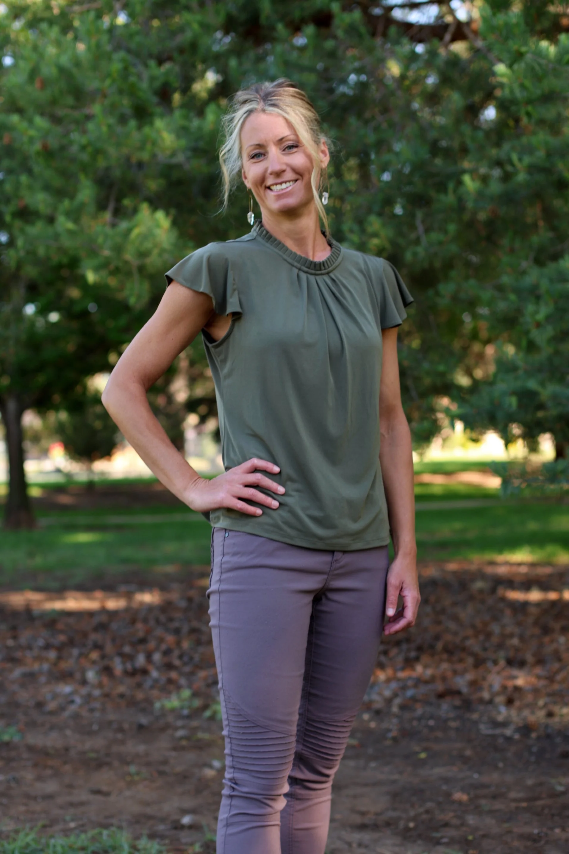 A woman with blond hair and blue eyes smiling outdoors near green pine trees, wearing a cream-colored long-sleeve top and gray pants.