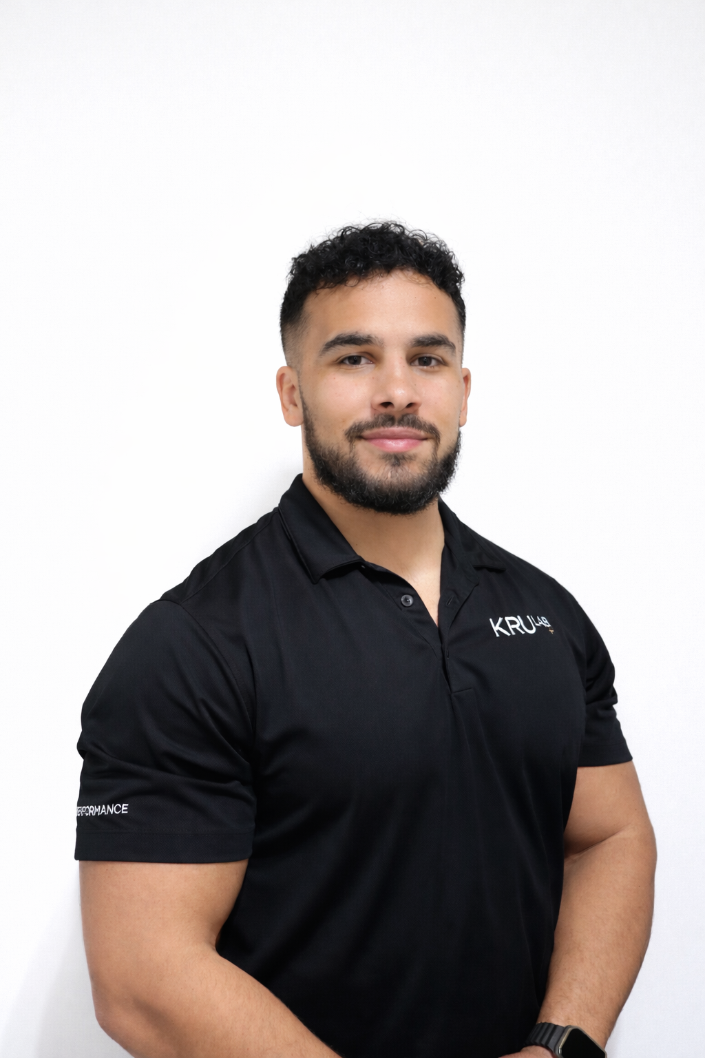 A young man with curly dark hair, beard, and mustache, wearing a black polo shirt with the words 'KRUA' and 'PERFORMANCE' on it, standing against a plain white background.