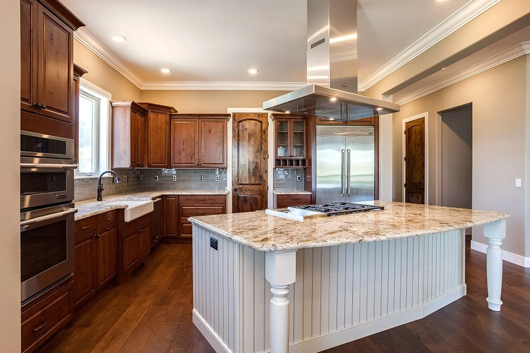 A rustic and warm kitchen with a stone top island and countertop, island range hood, stove in island, stainless steel fridge, brown matching floors, and white island base.