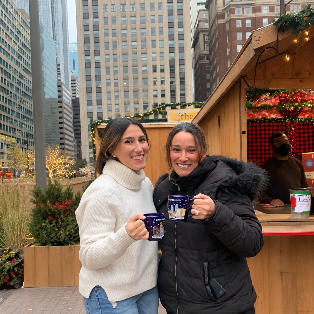 Two women smiling and holding holiday-themed mugs at an outdoor Christmas market in a city with tall buildings, decorated with Christmas lights and greenery.