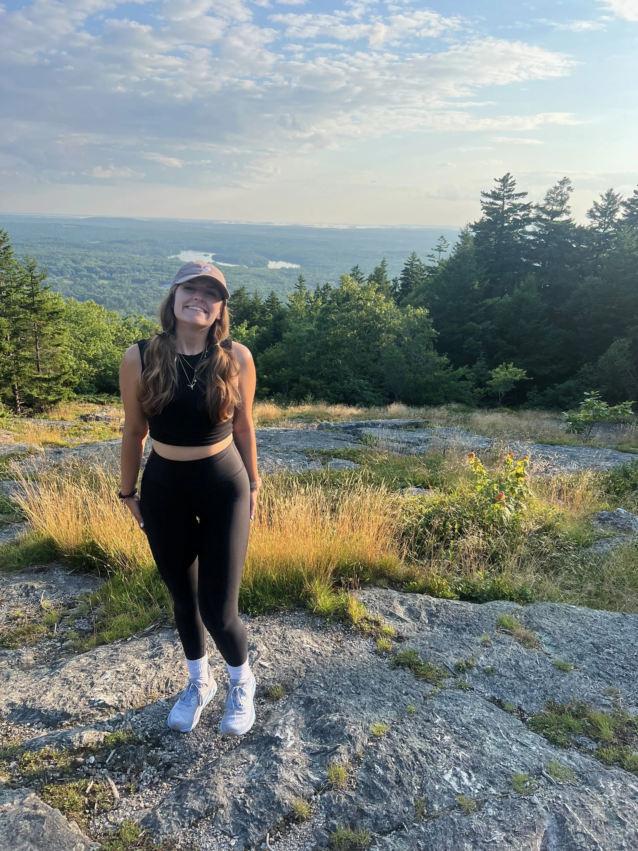 A young woman in black athletic clothing, a beige cap, and white sneakers, standing outdoors on rocky terrain with greenery and trees, smiling at the camera with a scenic view of trees and a river in the background during the daytime.