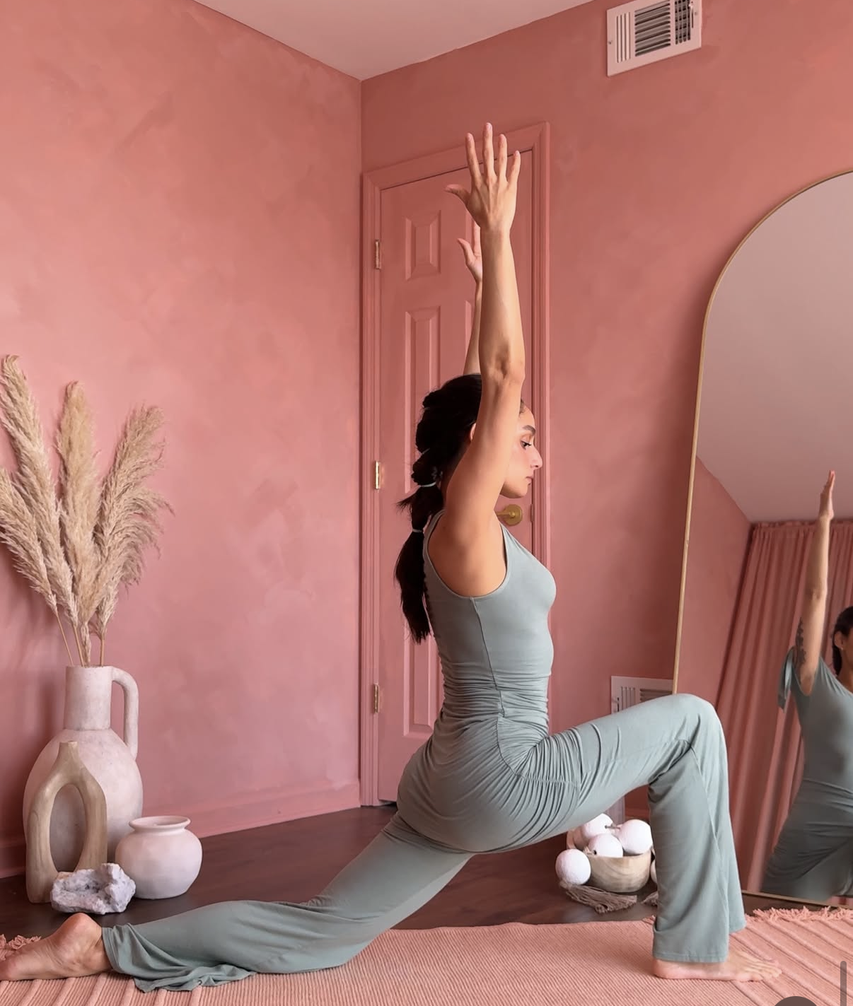 A woman practicing yoga indoors in a pink-themed room, performing a lunge stretch with arms raised, reflected in a mirror.