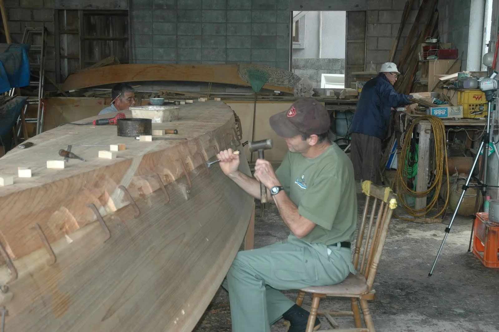 Douglas Brooks using a chisel and hammer to work on a traditional wooden boat hull in a workshop, with a Japanese master boatbuilder in the background.