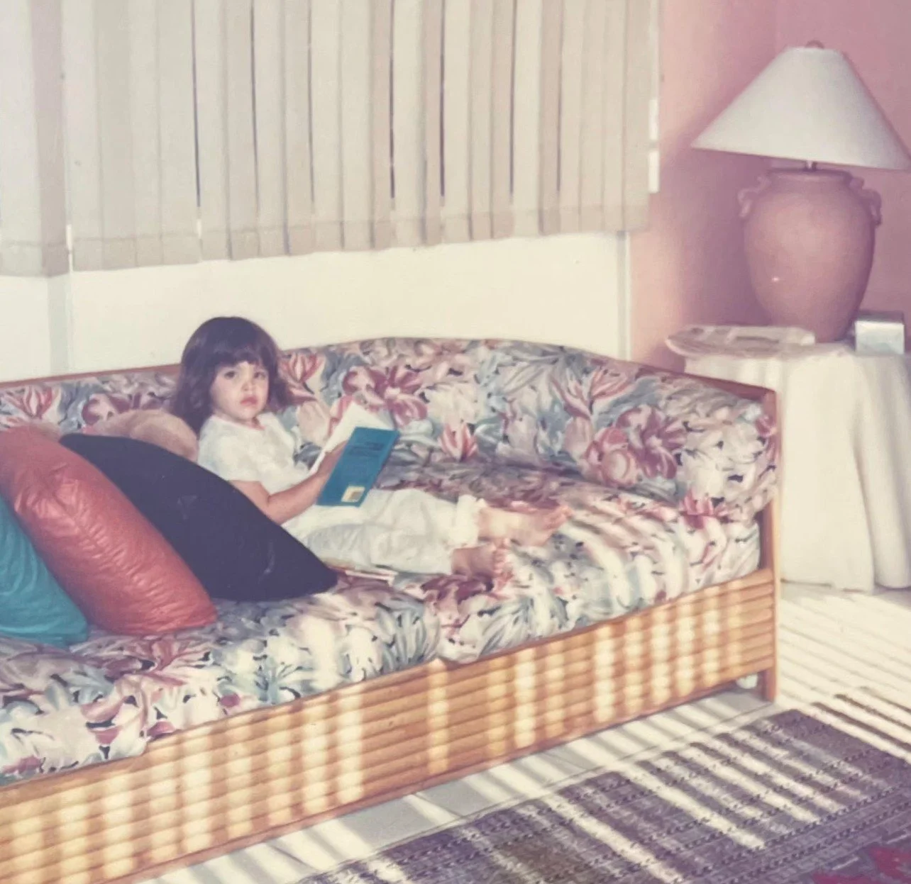 A young girl with dark hair and bangs is sitting on a floral-patterned couch, holding a book and looking towards the camera. The room has pink walls, beige blinds, a large table lamp on a side table with a stack of magazines, and a striped rug on the floor.
