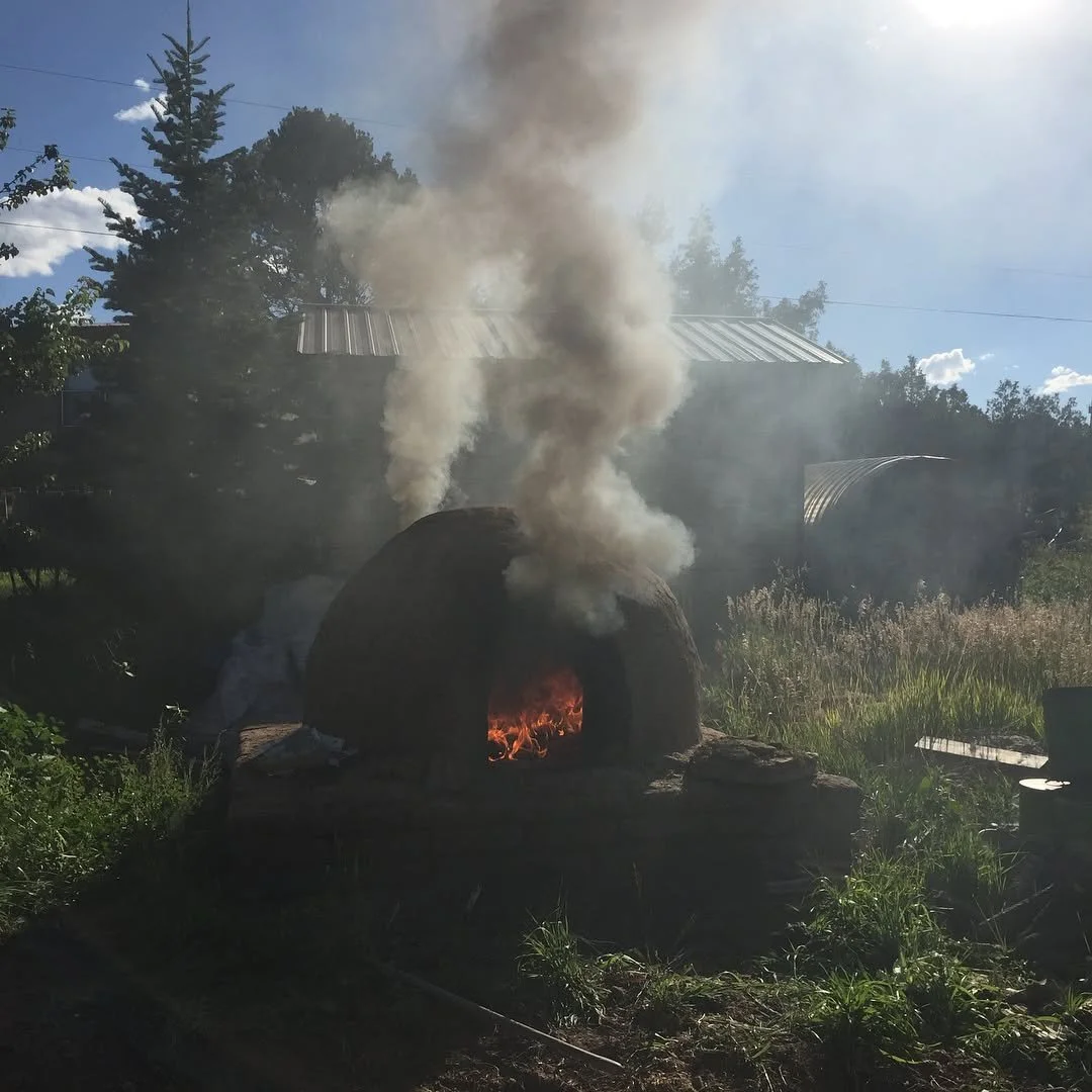 A wood-fired outdoor oven (or horno) with smoke and flames inside, emitting smoke clouds into the sky outdoors on a sunny day, surrounded by lush greenery.