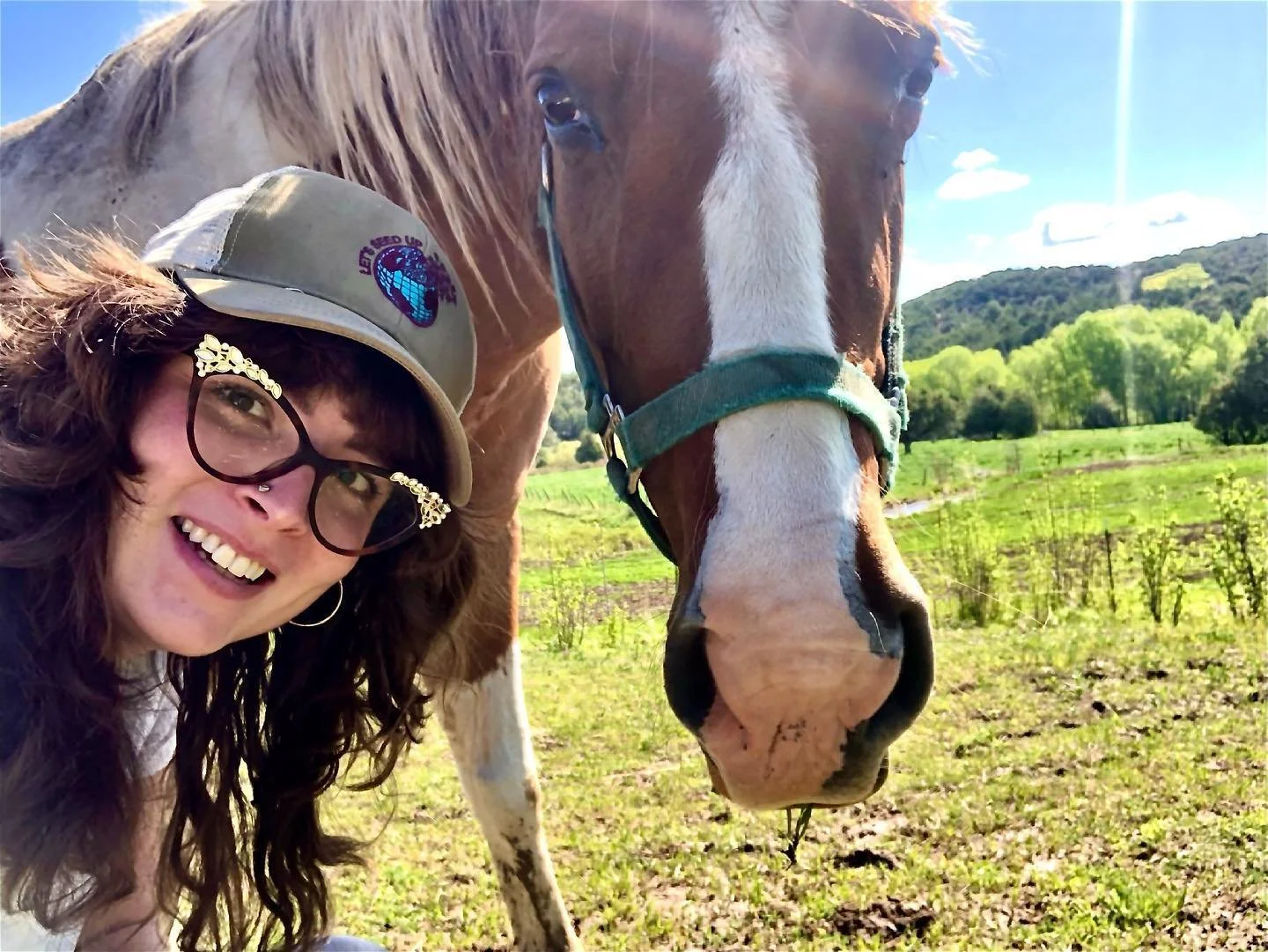 A smiling woman with long brown hair, glasses, and a beige cap with a blue logo, taking a selfie with a large, brown and white horse in a green field, sunny sky, and hills in the background.