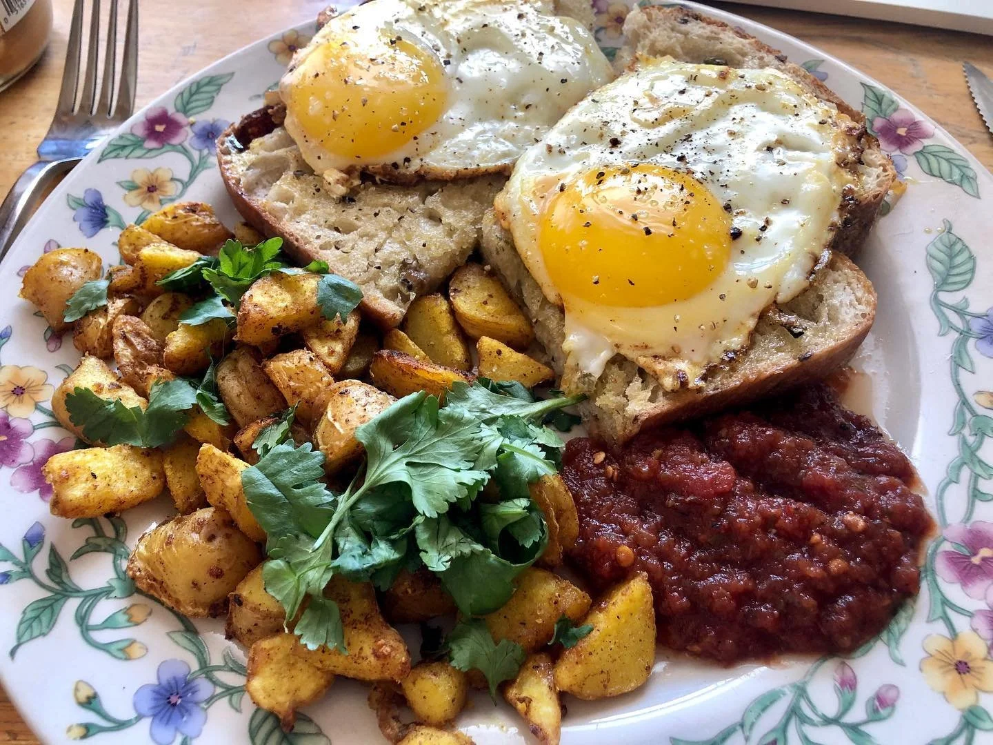 A plate of traditional breakfast food including two fried eggs, roasted potatoes with herbs, crispy fried bread, and red chili sauce, all served on a floral-patterned plate.