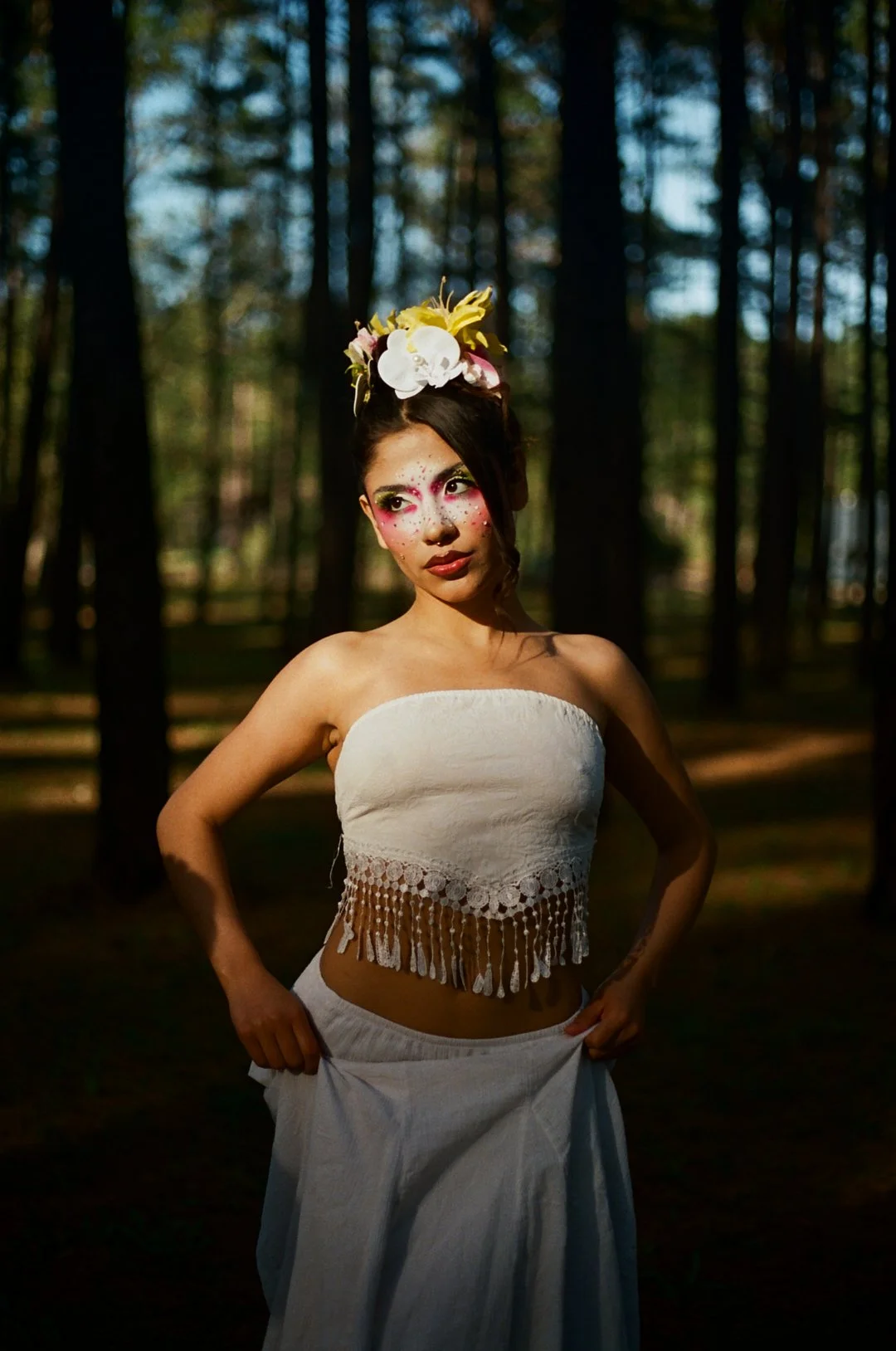A young woman standing in a forest with sunlit trees in the background, wearing a strapless white top with fringe details and a matching skirt, with her hands holding the skirt and decorated with floral accessories and artistic makeup.