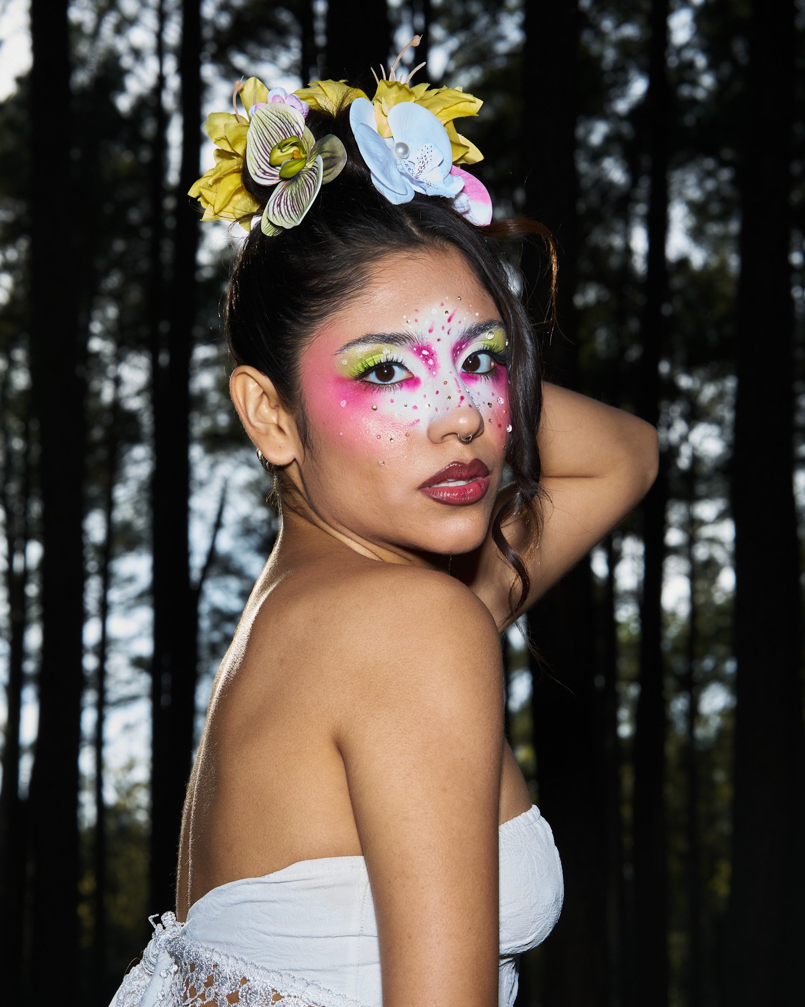 A woman with colorful makeup, earrings, and a headpiece made of flowers poses outdoors in front of a dark forest.