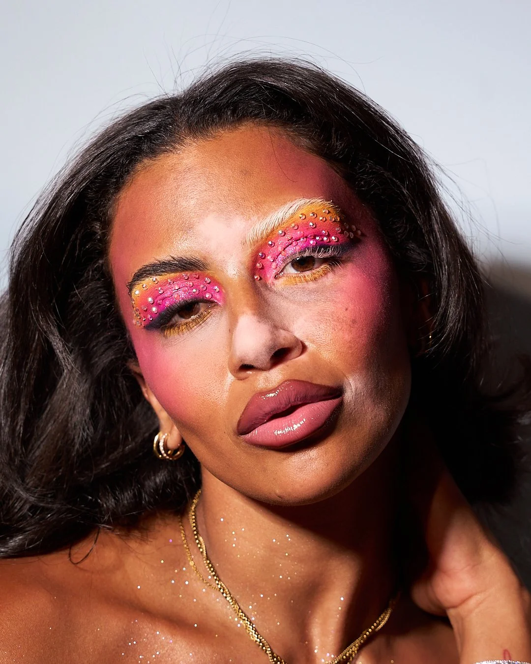 Close-up of a woman with vibrant pink eye makeup decorated with rhinestones, gold accents, glossy lips, dark wavy hair, gold earrings, and a glittery necklace.