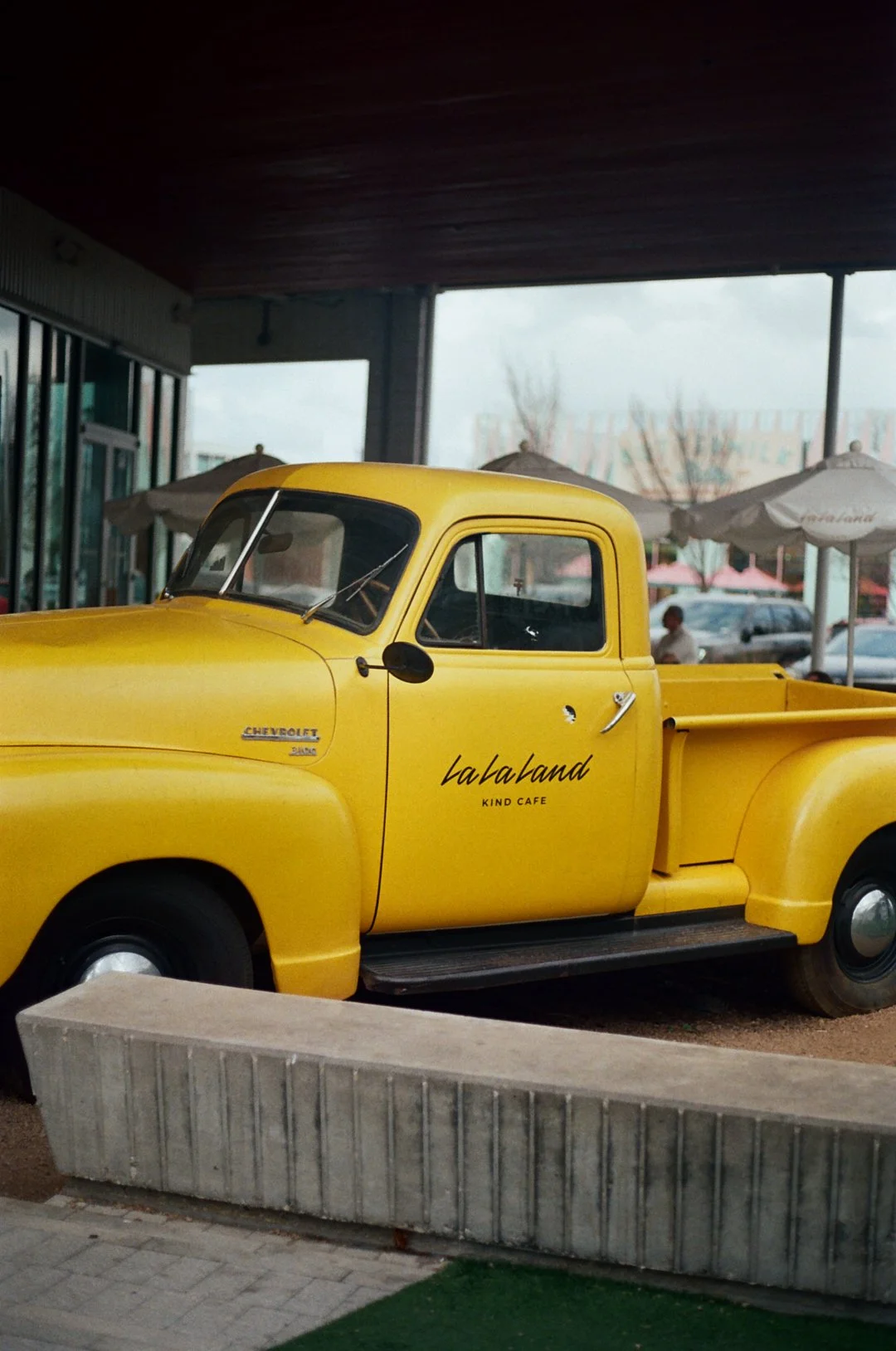 Yellow vintage Chevrolet truck with 'Lala Land' and 'Kind Cafe' written on the door, parked outside under a roof with umbrellas and people in the background.