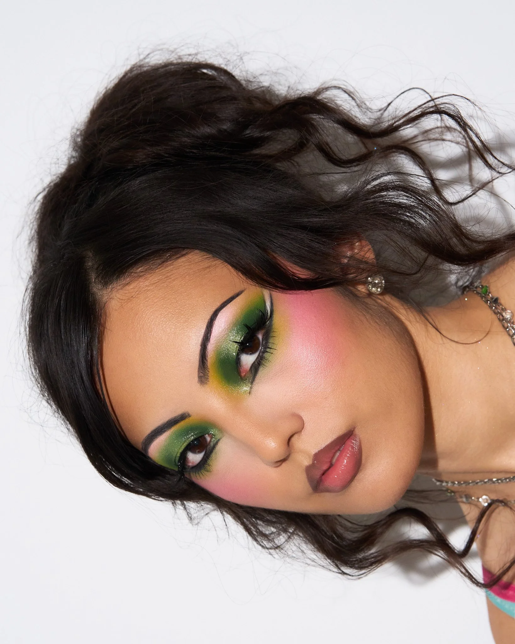 Close-up of a woman with vibrant green and pink makeup, black hair, and jewelry against a white background.