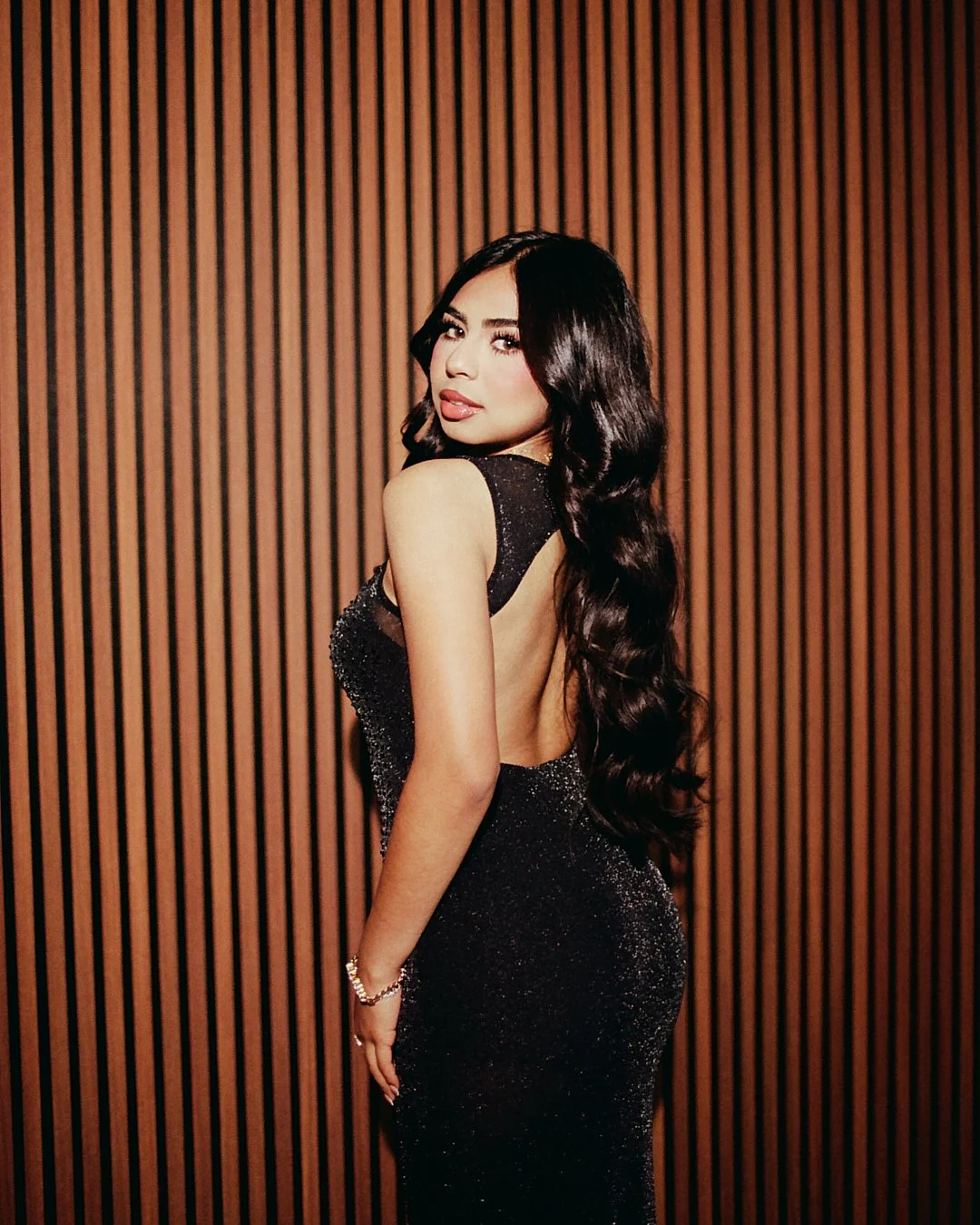 A woman with long wavy dark hair standing against a wooden slat wall, wearing a black sparkly backless gown and a bracelet.