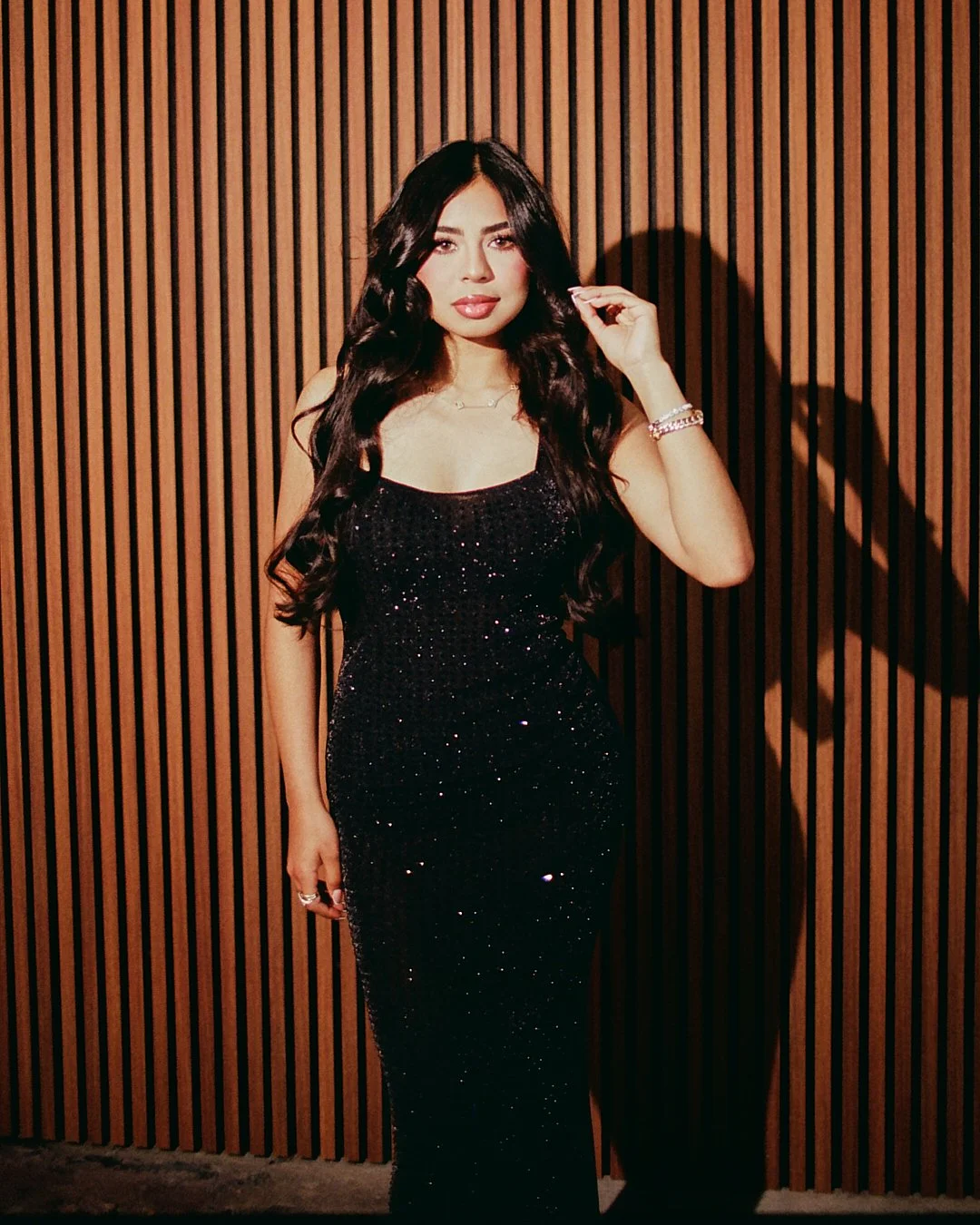A woman in a black, sparkly, floor-length gown with long black hair stands against a wooden slat wall. She has a bracelet and rings on her right hand and touches her hair.