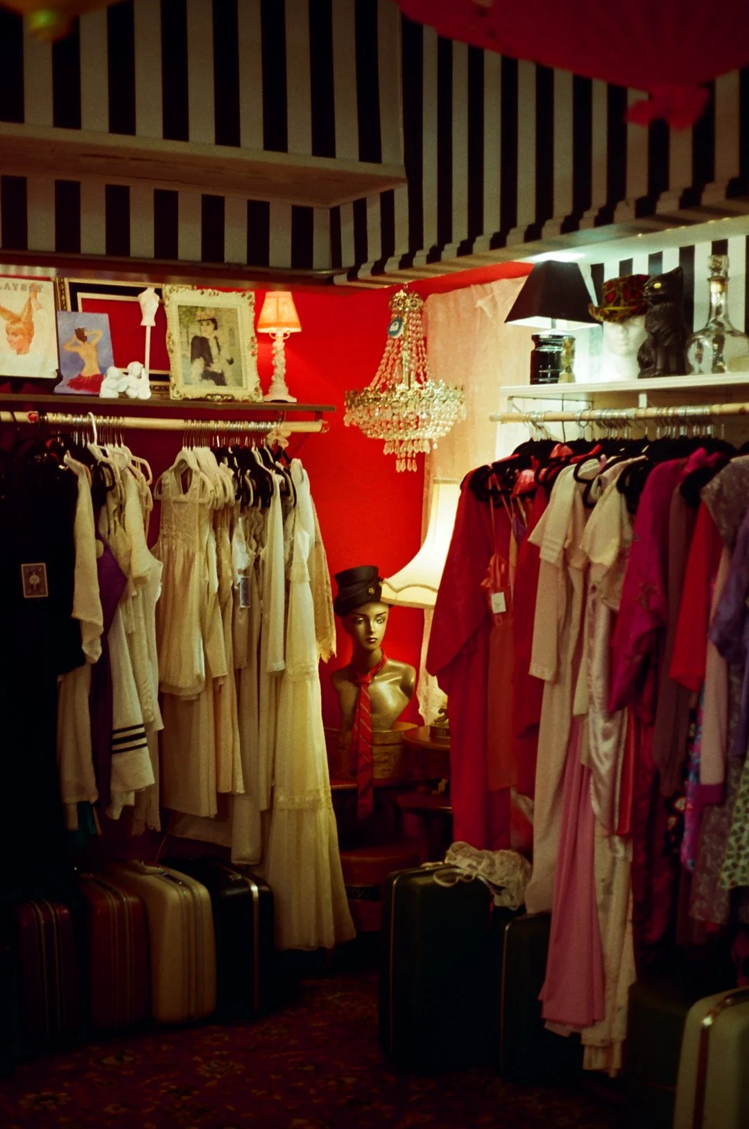 Clothing store with vintage dresses, a mannequin wearing a headscarf, and decorative items like picture frames, hats, lamps, and a chandelier.