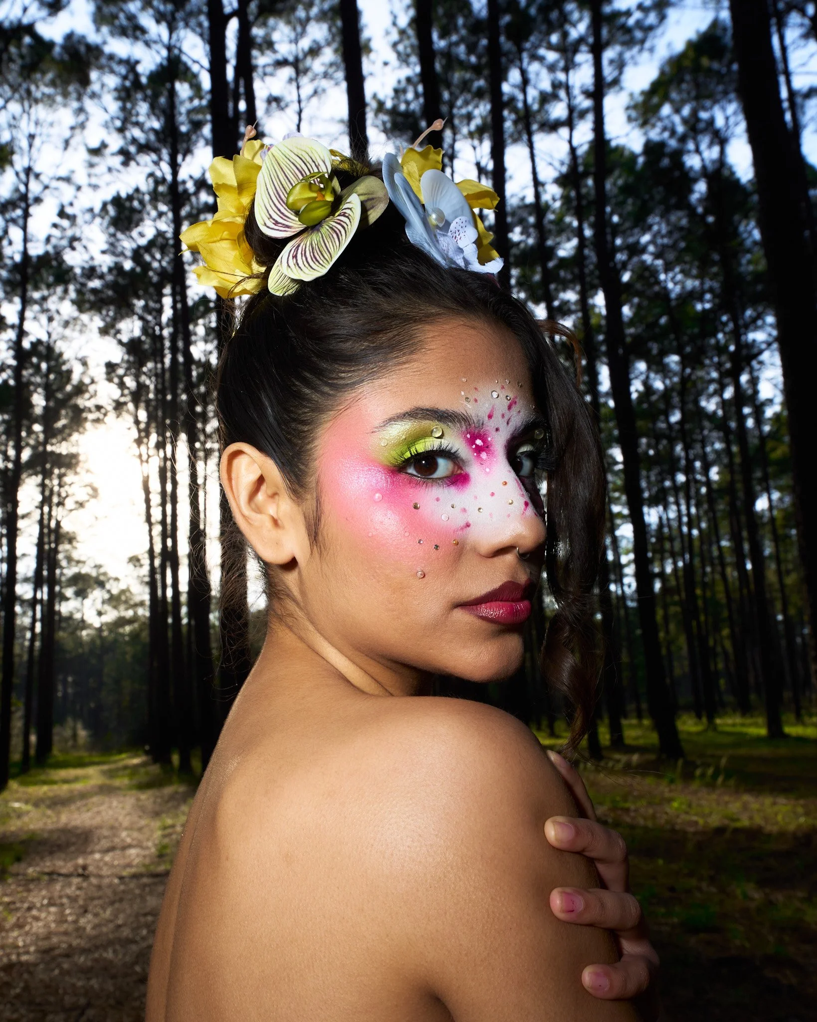 A woman with colorful makeup, decorative face gems, and floral headpiece in a forest setting.