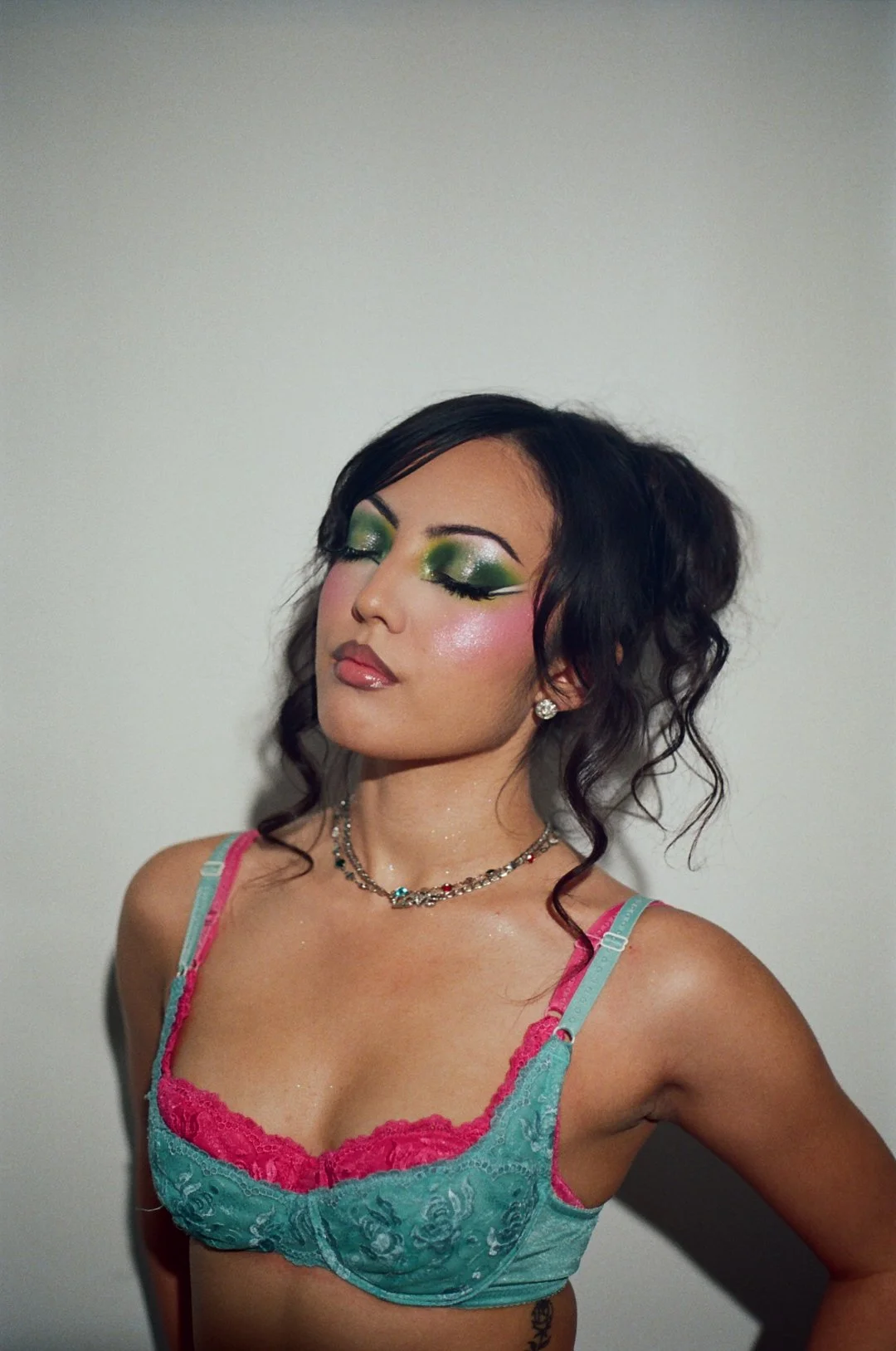 Woman with curly dark hair and green and pink makeup, wearing a teal and pink lace bra, and layered necklaces, standing against a plain background with eyes closed.
