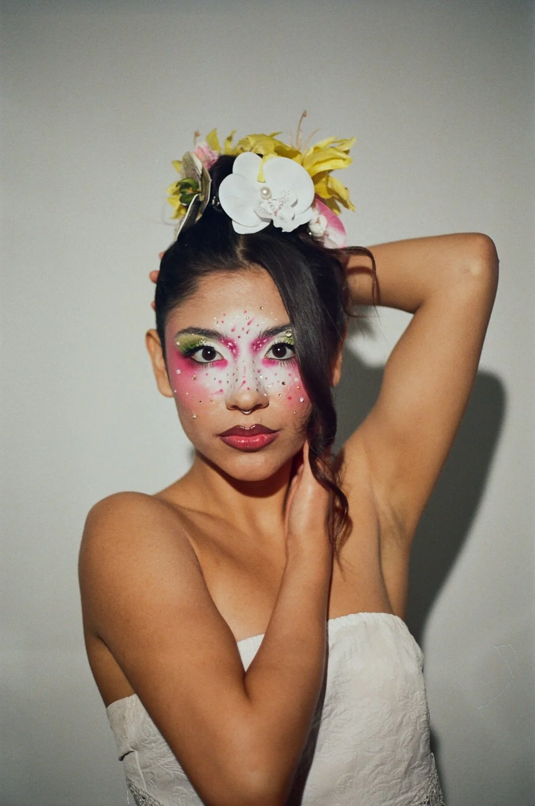 A woman with elaborate makeup resembling a butterfly, with pink, white, and green colors, adorned with rhinestones. She has a large floral headpiece with white, yellow, and pink flowers. She is wrapped in a white fabric and poses with one hand behind her head and the other touching her neck against a plain background.