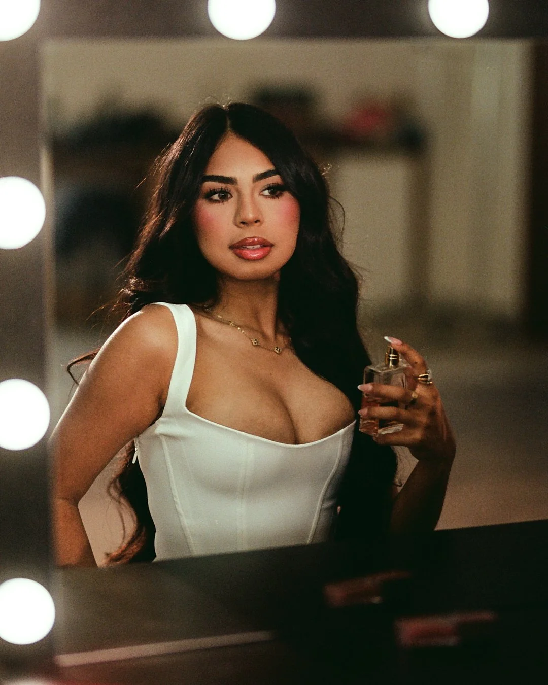 A woman with long dark hair, wearing a white dress with a deep neckline, standing in front of a mirror with lights around it, holding a bottle of perfume, in a dressing room or backstage area.