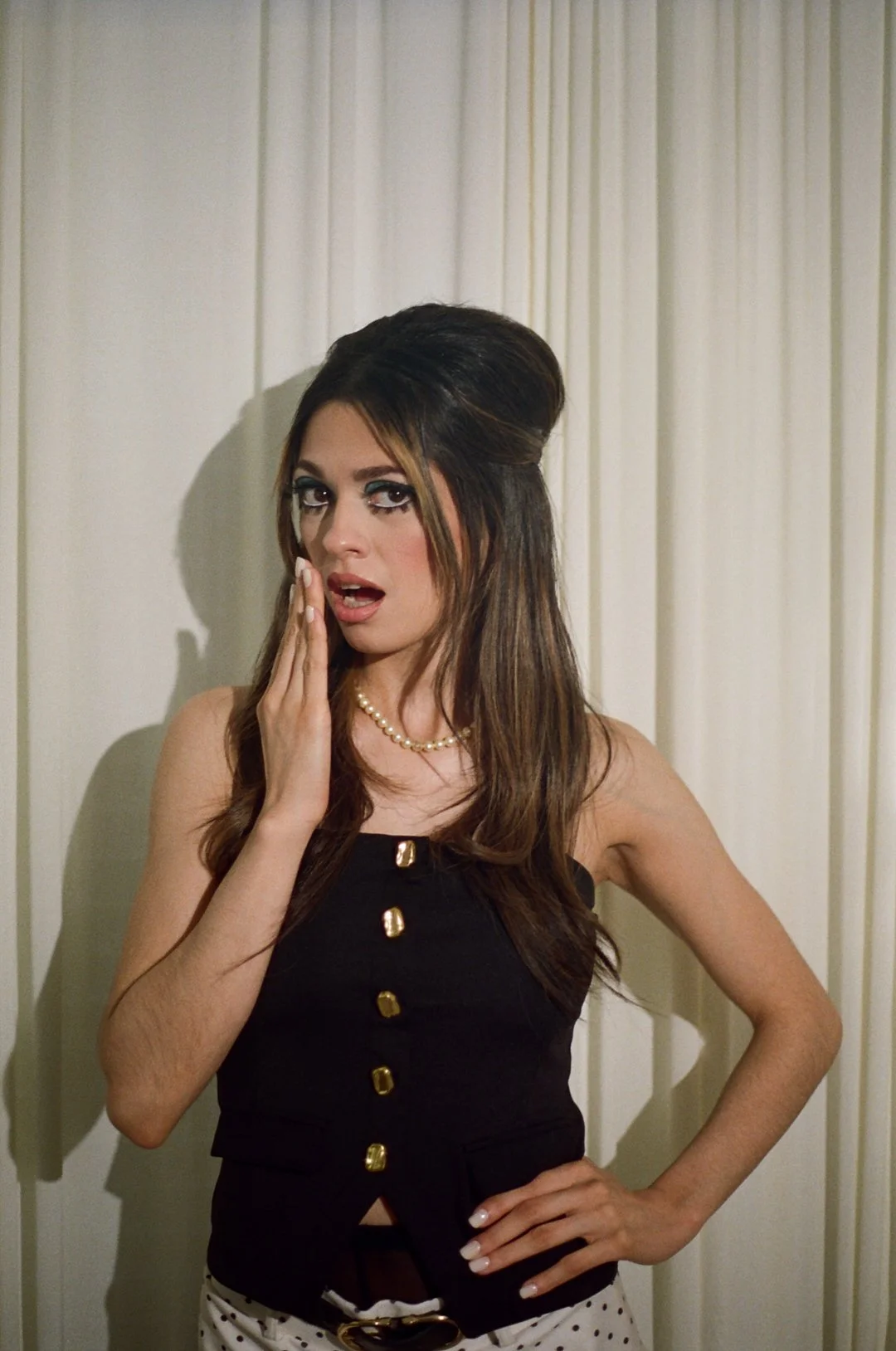 A woman with long brown hair styled in a retro do, wearing a black sleeveless top with gold buttons, a pearl necklace, and white polka dot pants, standing in front of a cream-colored curtain, with a surprised expression, hand on her cheek.