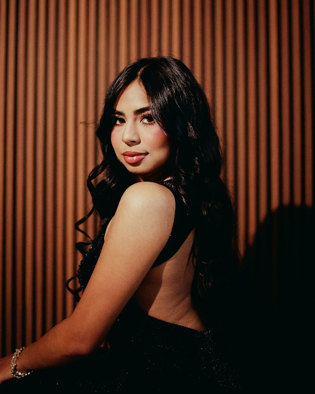 A woman with long dark hair and makeup posing in front of a wooden panel background, wearing a black backless dress with sparkling details, smiling softly at the camera.
